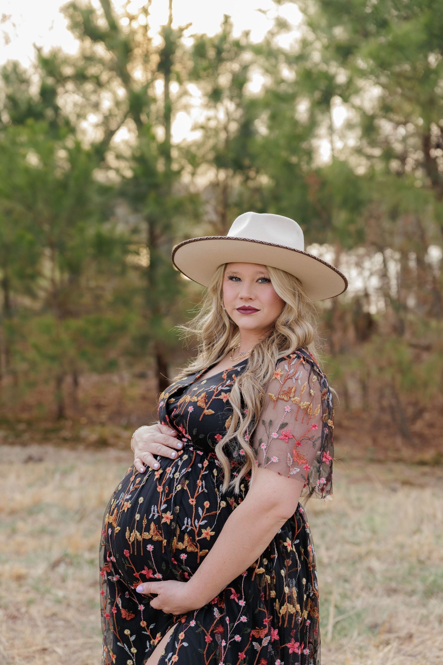 Pregnant woman in floral dress and wide-brim hat cradling baby bump outdoors in a wooded setting.