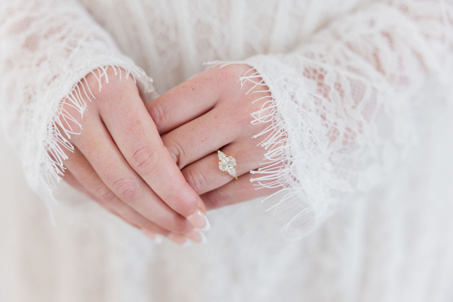 Close-up of a bride's hands wearing a sparkling diamond engagement ring, dressed in elegant white lace wedding gown sleeves.