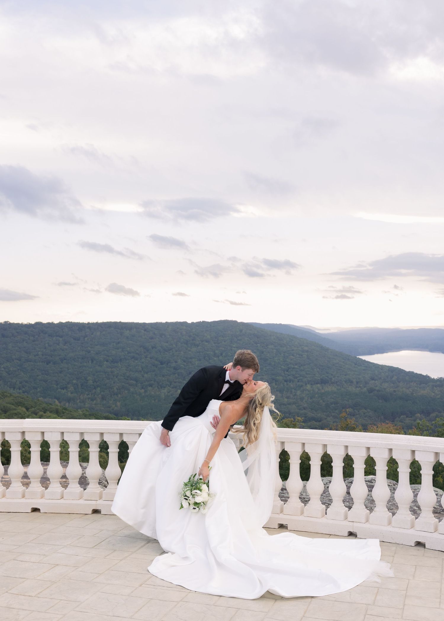 Bride and groom share a romantic dip kiss on a balcony overlooking lush mountains and a serene lake at sunset.