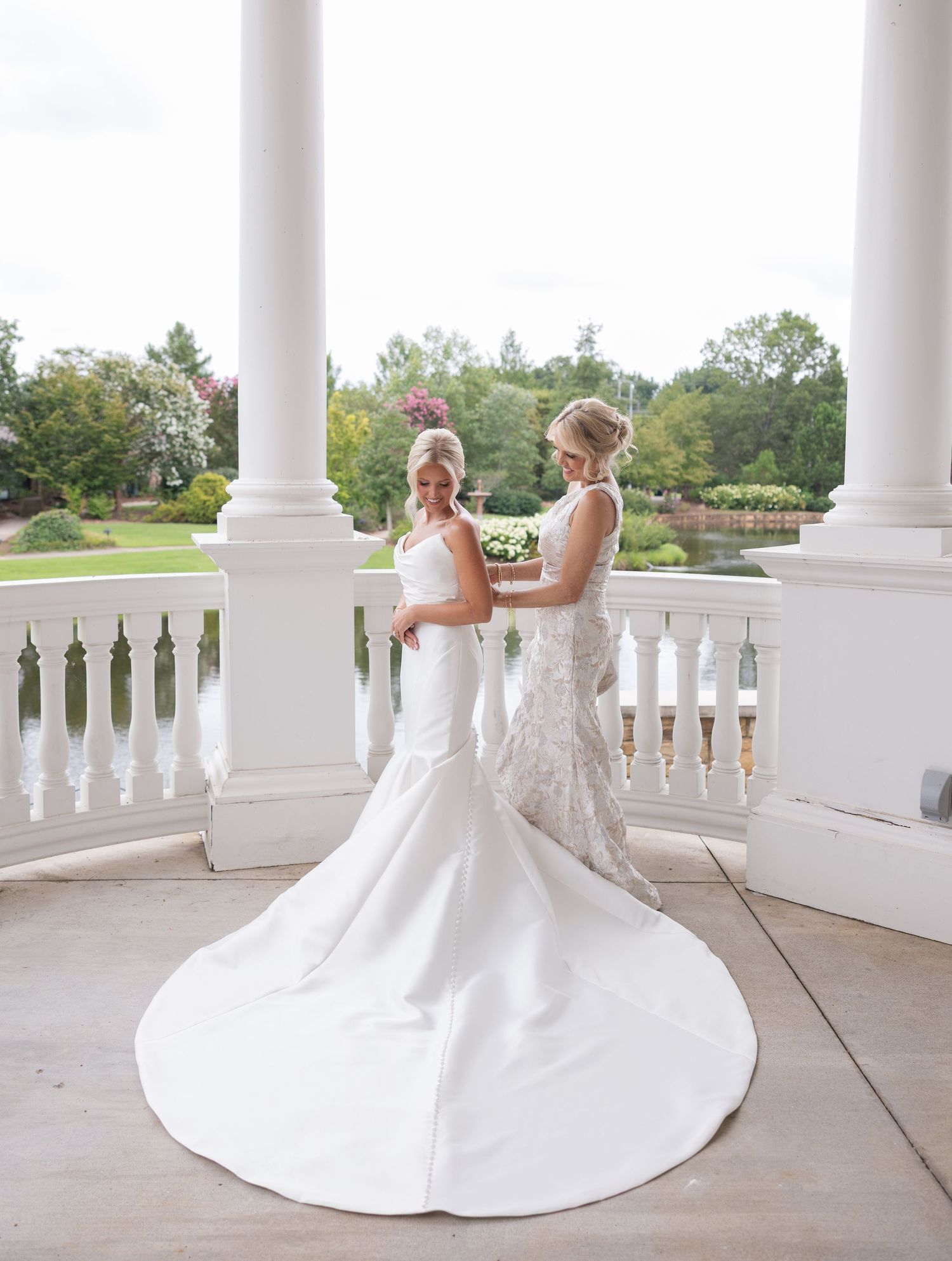 Two brides in elegant white wedding gowns pose on a grand columned terrace surrounded by lush gardens.