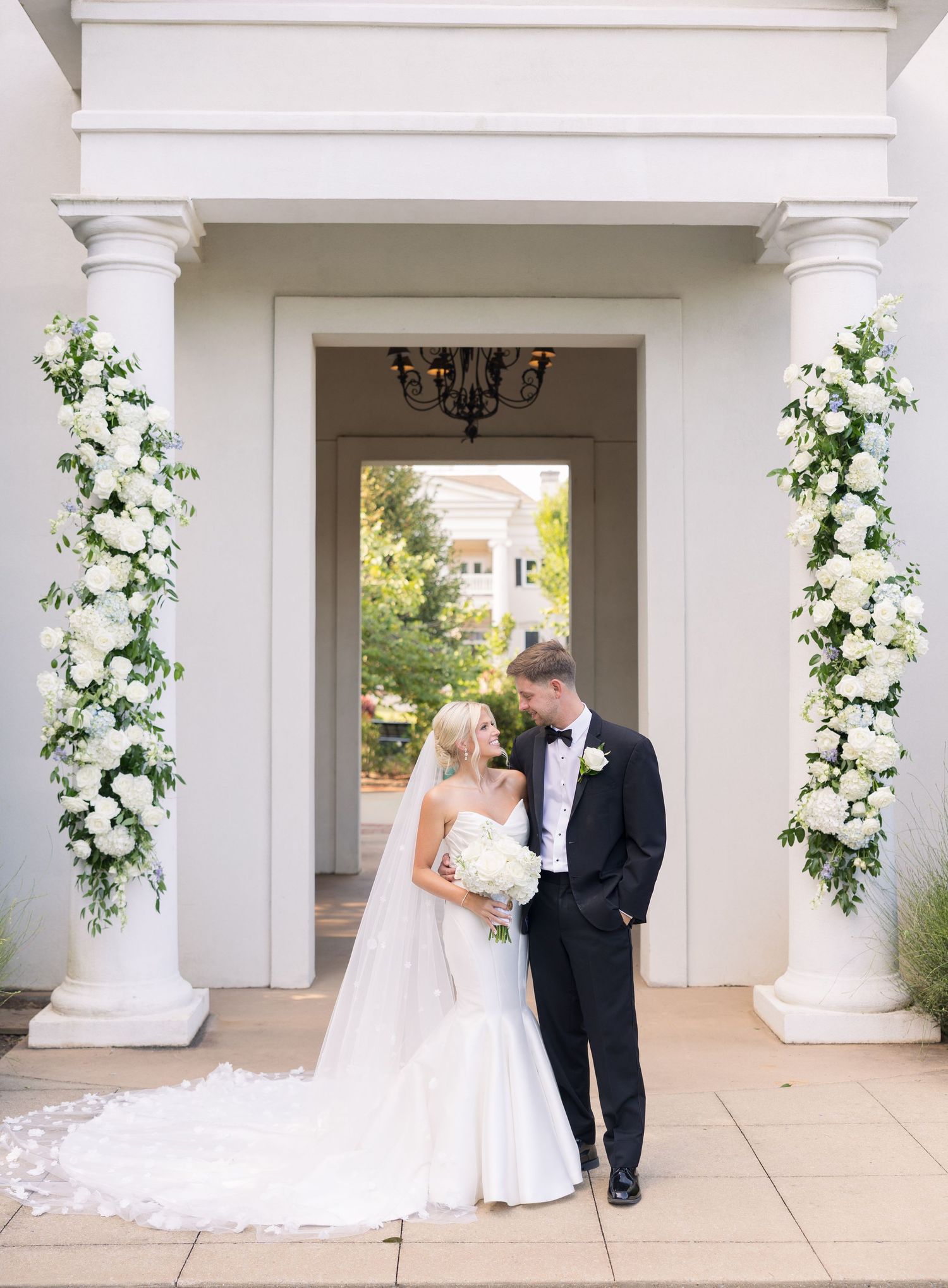 Bride and groom share a romantic moment between white columns adorned with lush floral arrangements at elegant wedding venue.