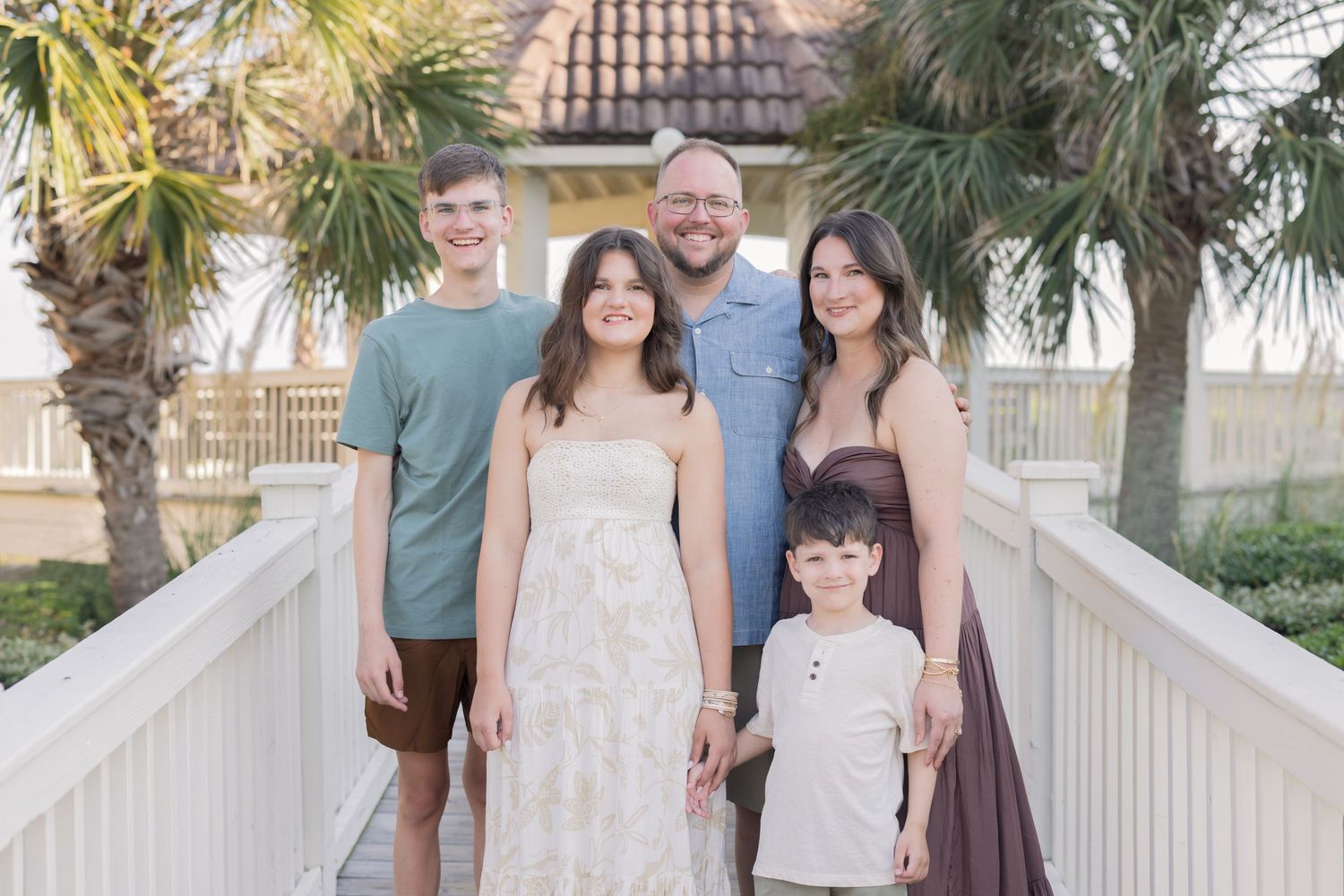 Family of five smiling together on a wooden bridge surrounded by palm trees in a sunny coastal setting.