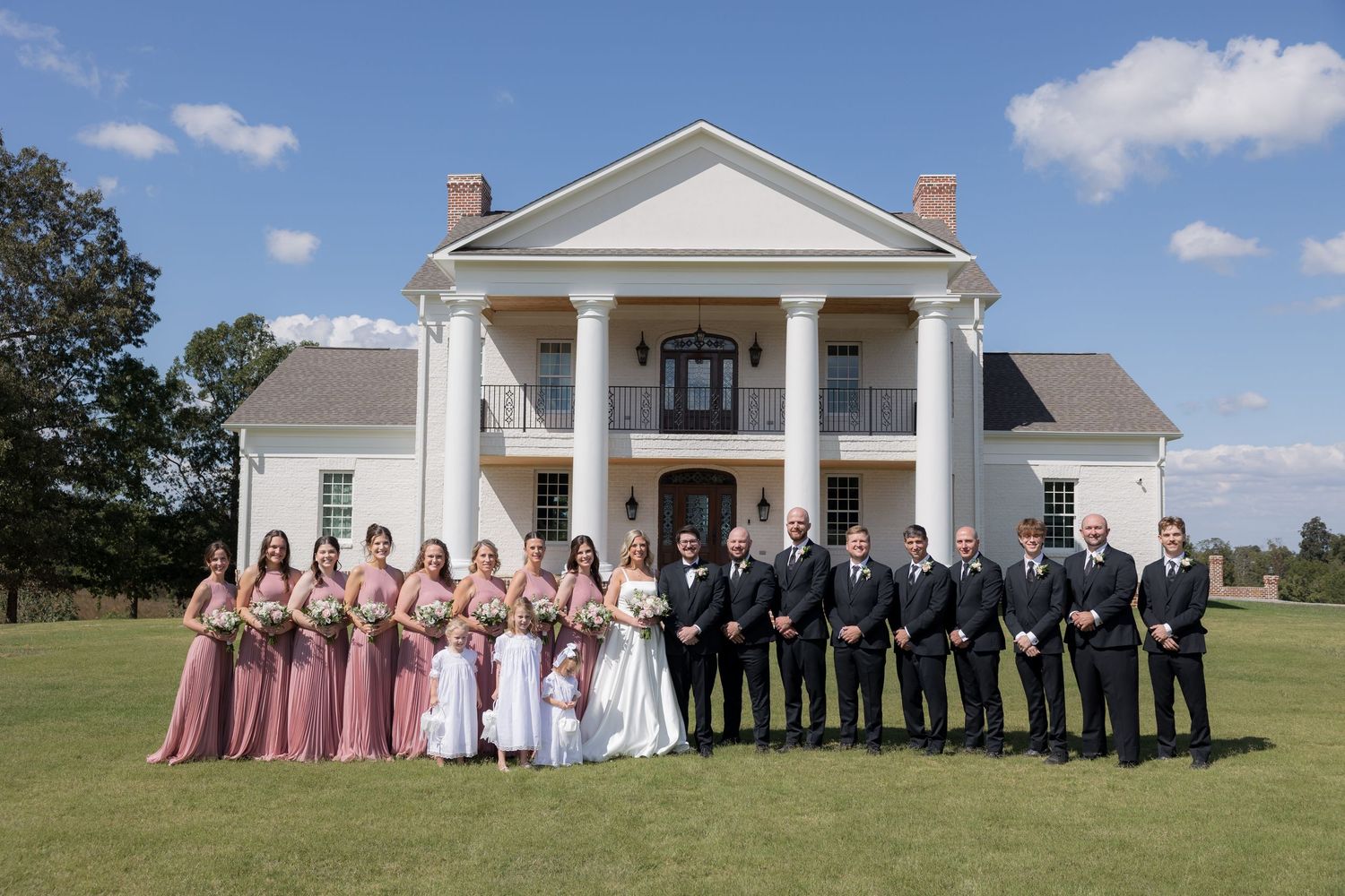 Wedding party posed in front of elegant white colonial venue with tall columns on sunny day.