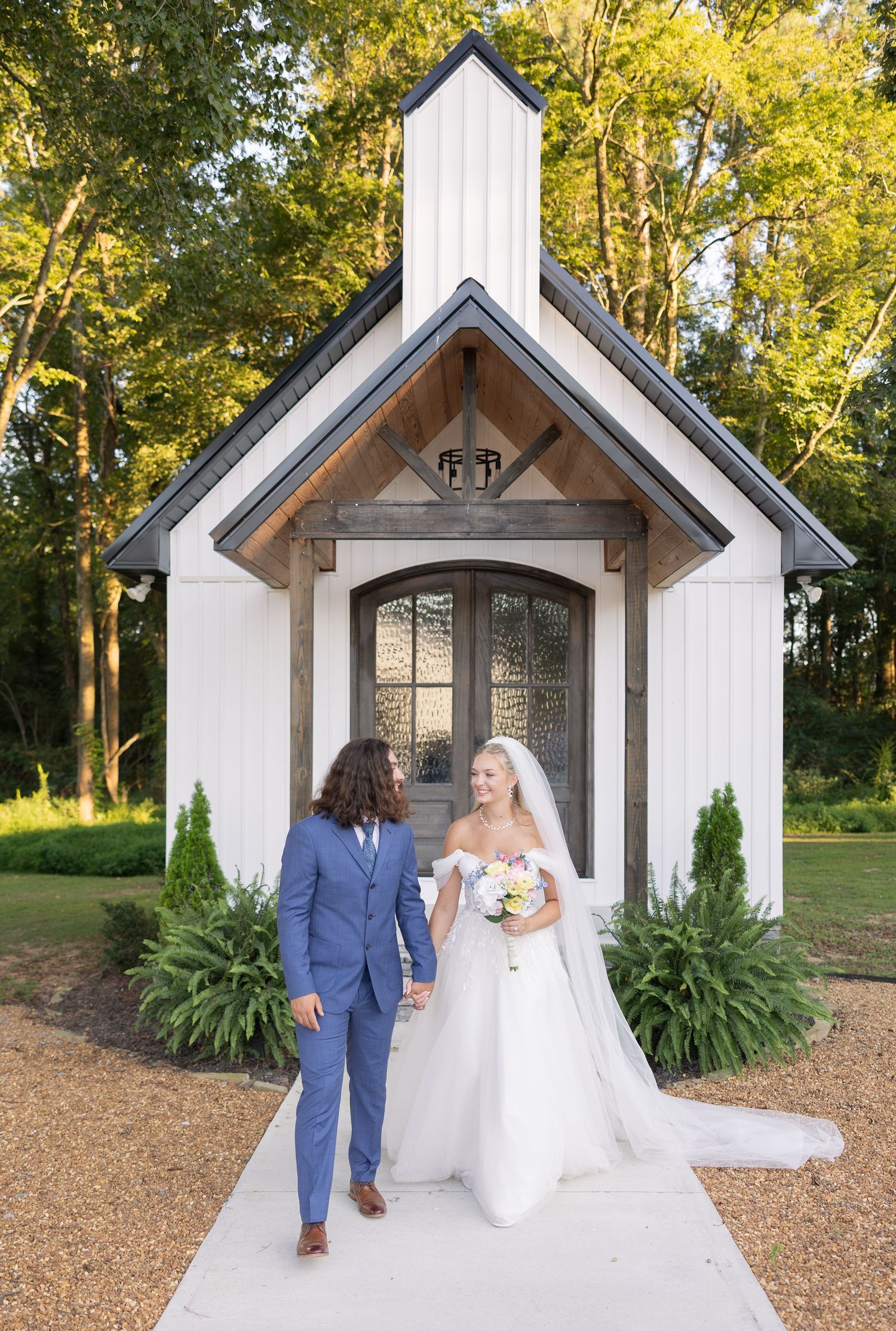 Bride and groom holding hands in front of a white chapel with arched doors, surrounded by lush greenery.