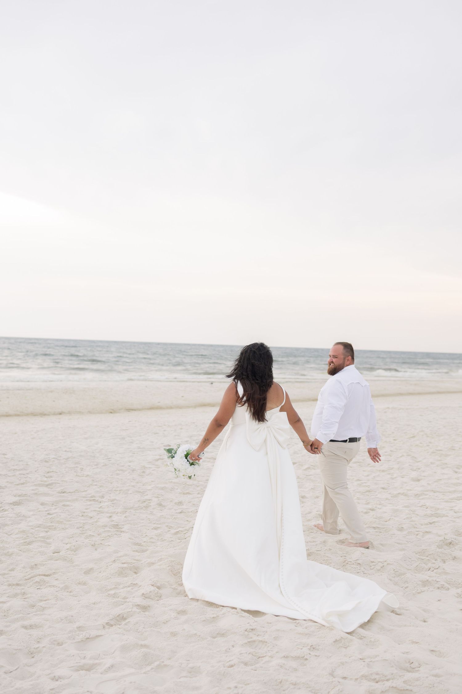 Bride in white gown and groom in white shirt walk hand-in-hand on a sandy beach at dusk.