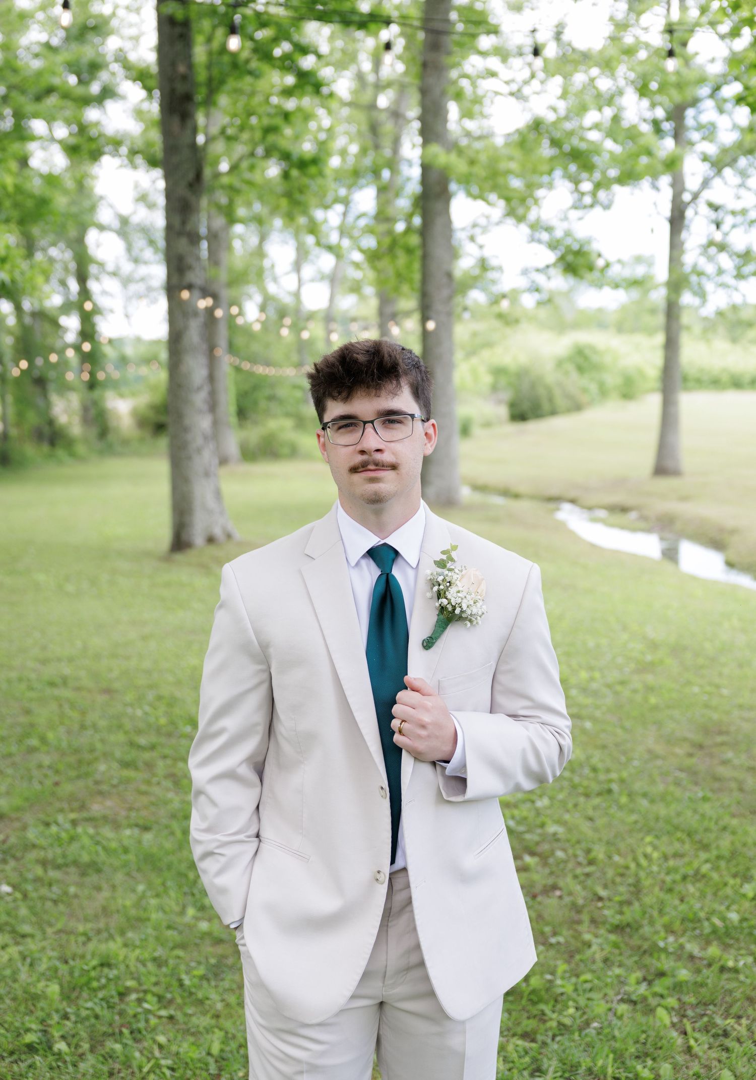 Young groom in beige suit with teal tie and boutonniere poses outdoors near trees and string lights.