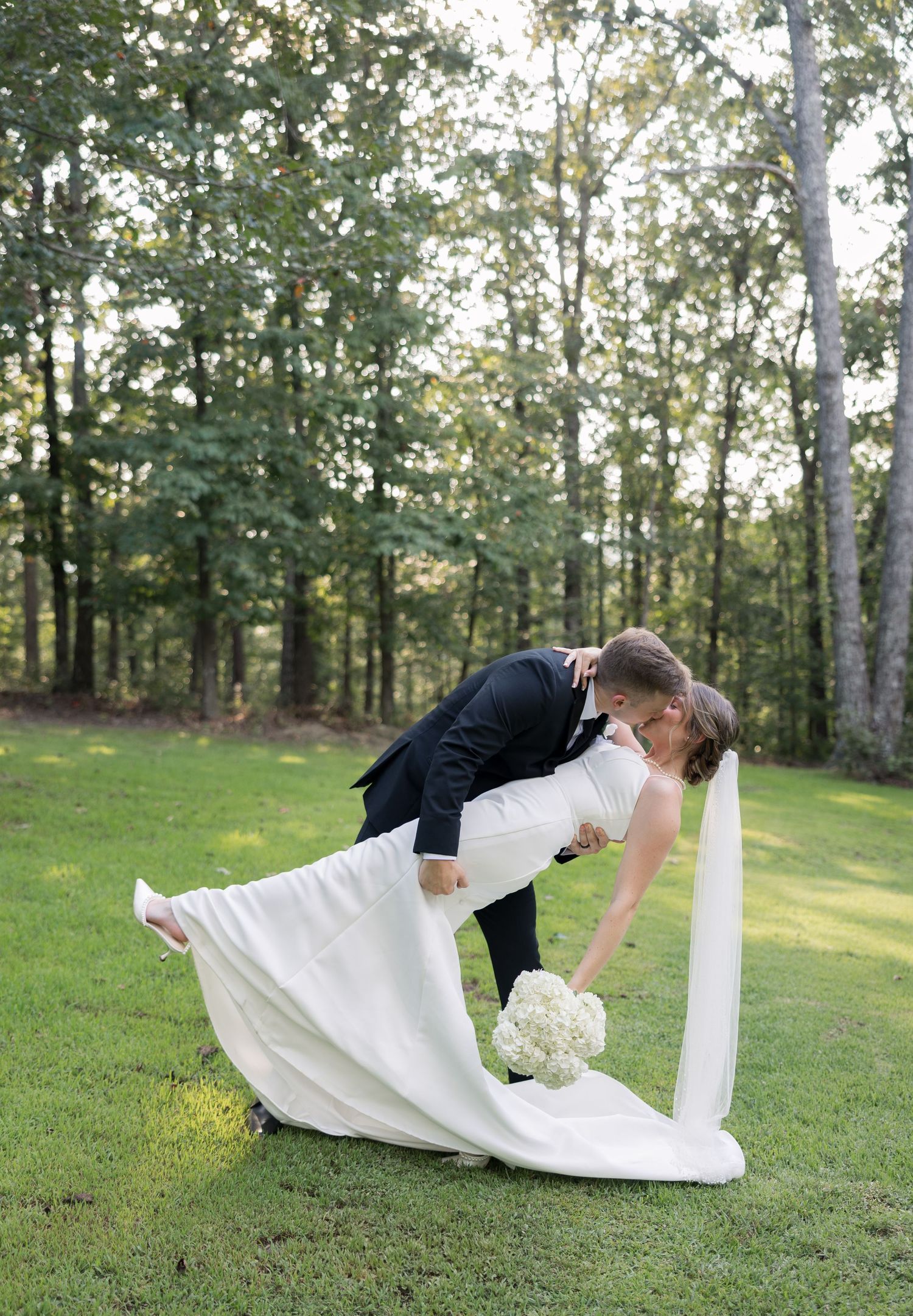 Groom dips bride in white gown and bouquet on green lawn with forest backdrop in romantic wedding photo.
