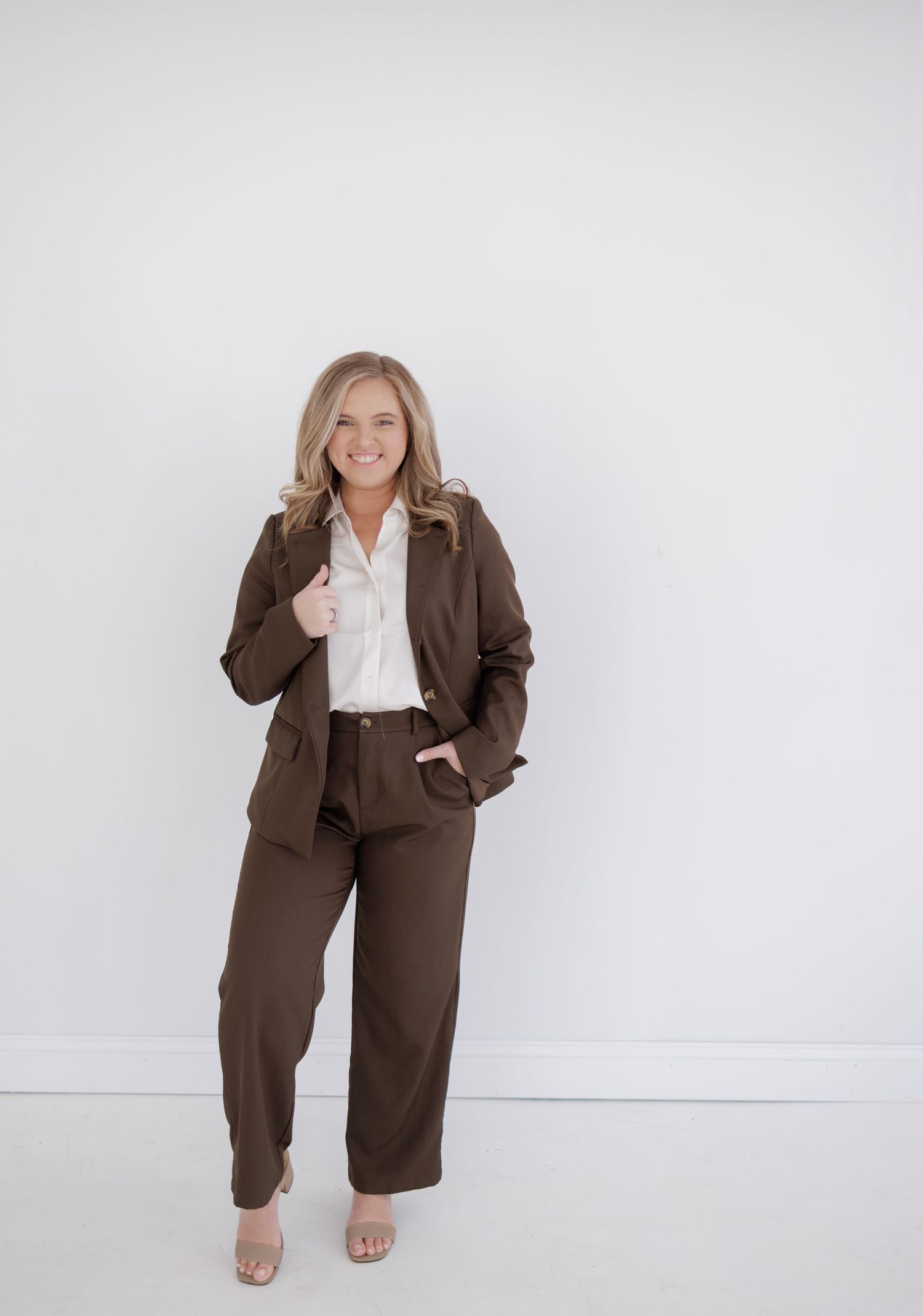 Woman in brown blazer and trousers with white blouse, smiling and giving thumbs up against white background.