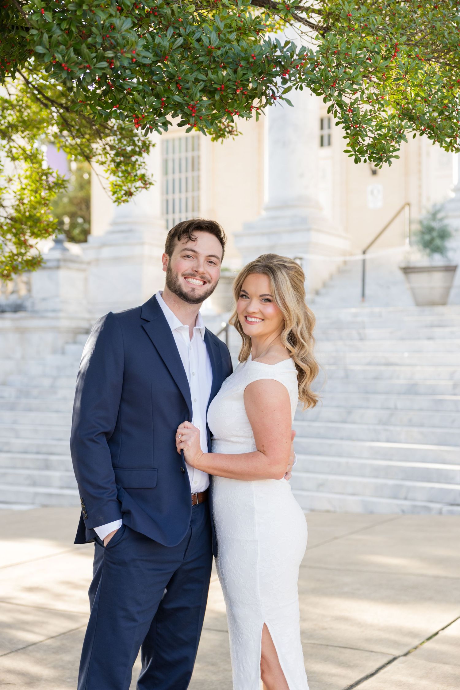 Smiling couple in wedding attire posing outdoors near classic white building steps under a berry tree.
