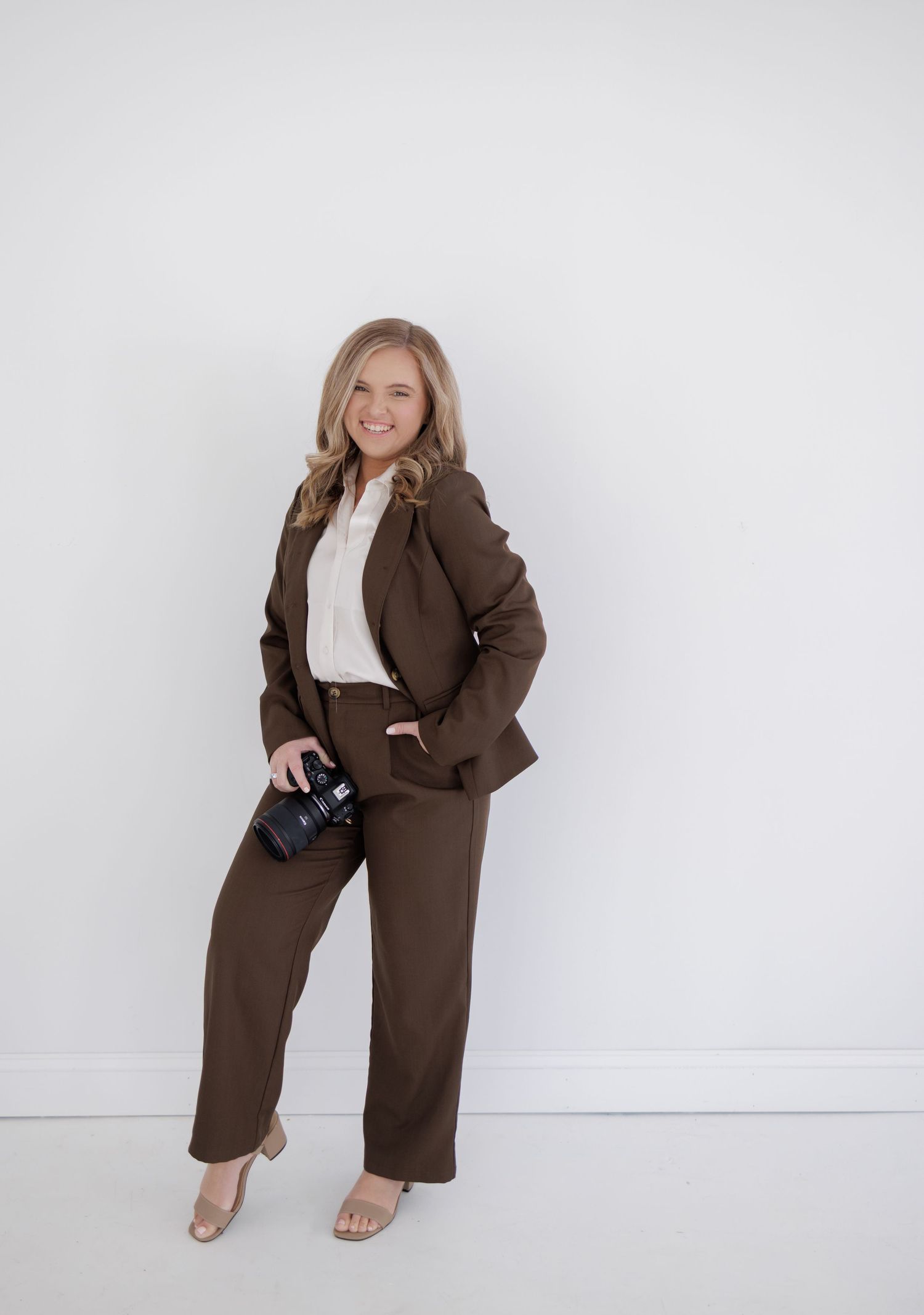 Confident blonde woman in brown pantsuit holds camera against a clean white background.