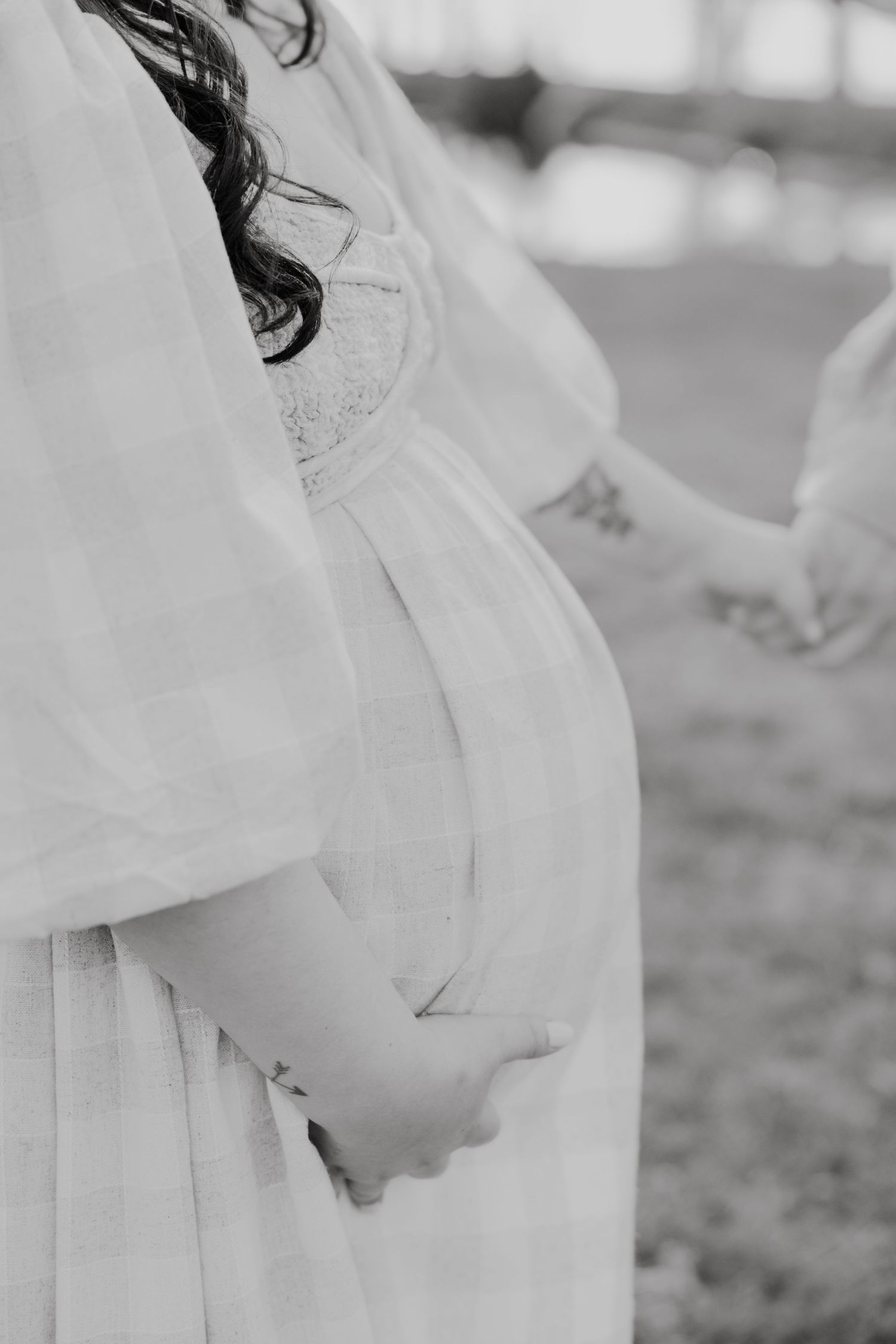 Black and white maternity photo of pregnant woman in white dress cradling baby bump while holding hands outdoors.