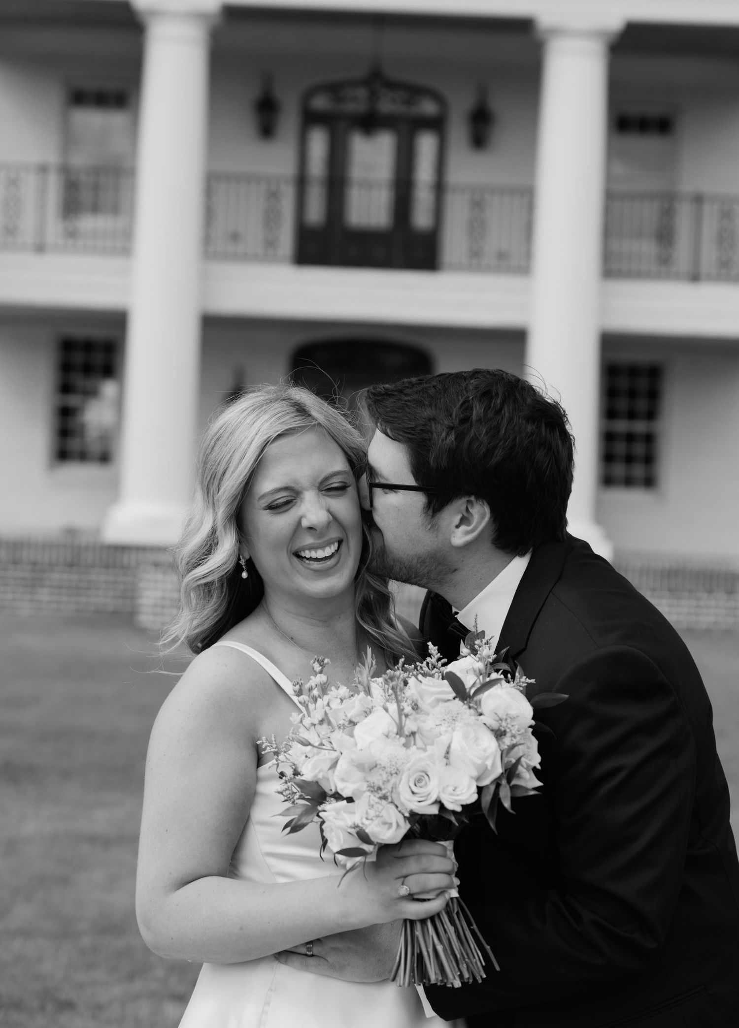 Black and white wedding photo of smiling bride holding bouquet as groom kisses her cheek before a grand plantation home.