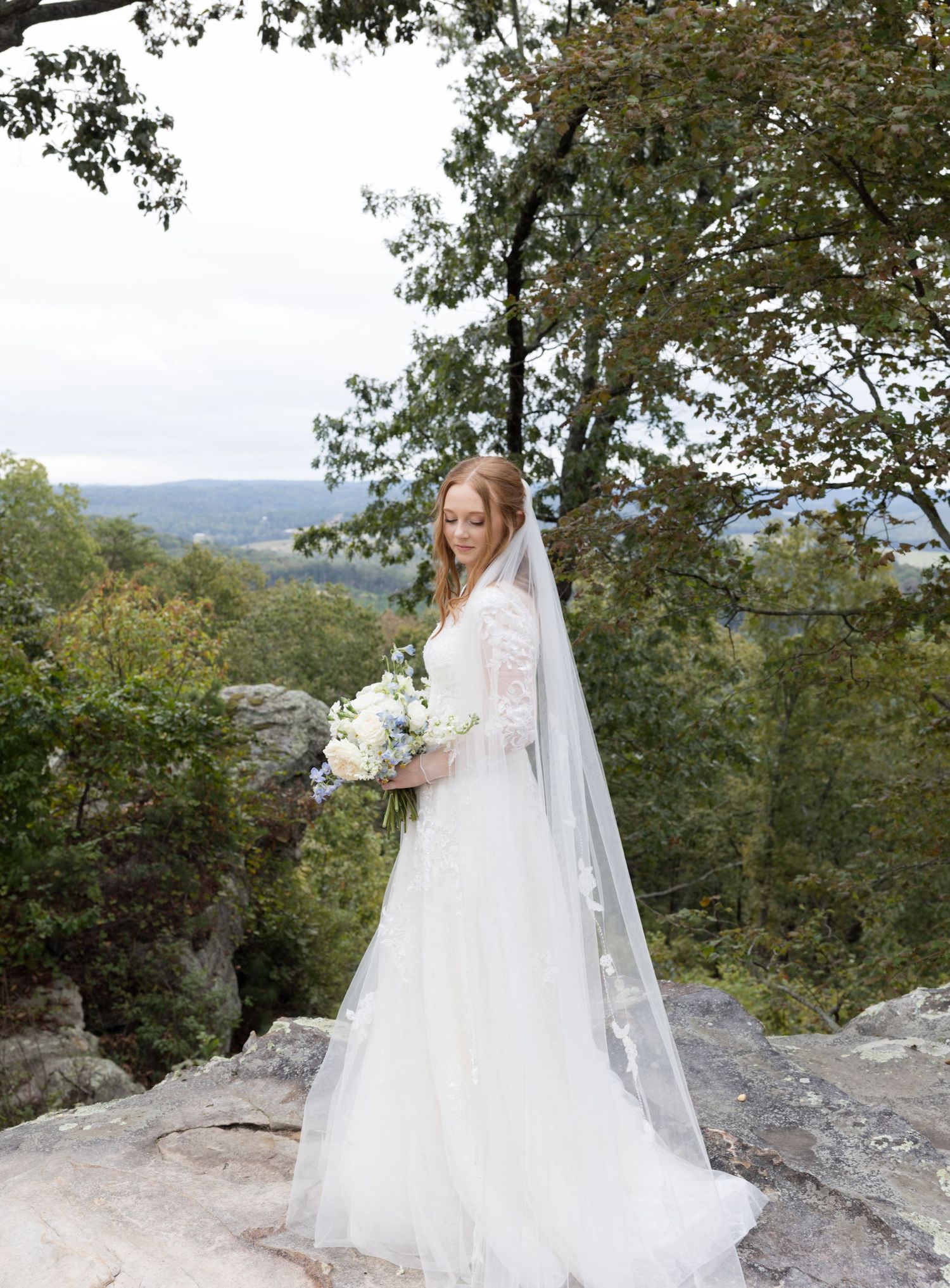 Bride in white gown and veil holds bouquet on rocky overlook surrounded by lush green trees.
