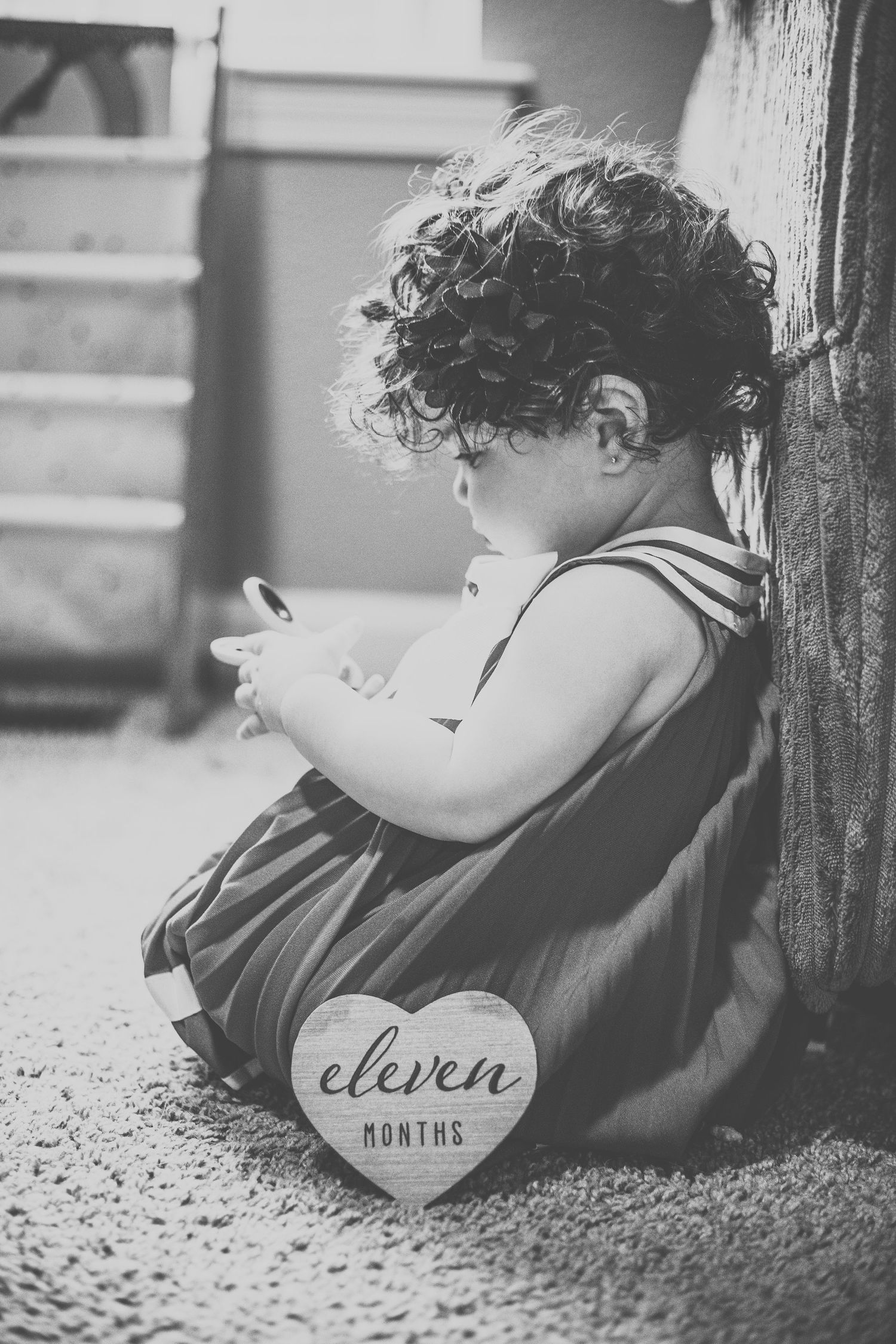 Black and white photo of an eleven-month-old baby with curly hair sitting against a tree trunk.