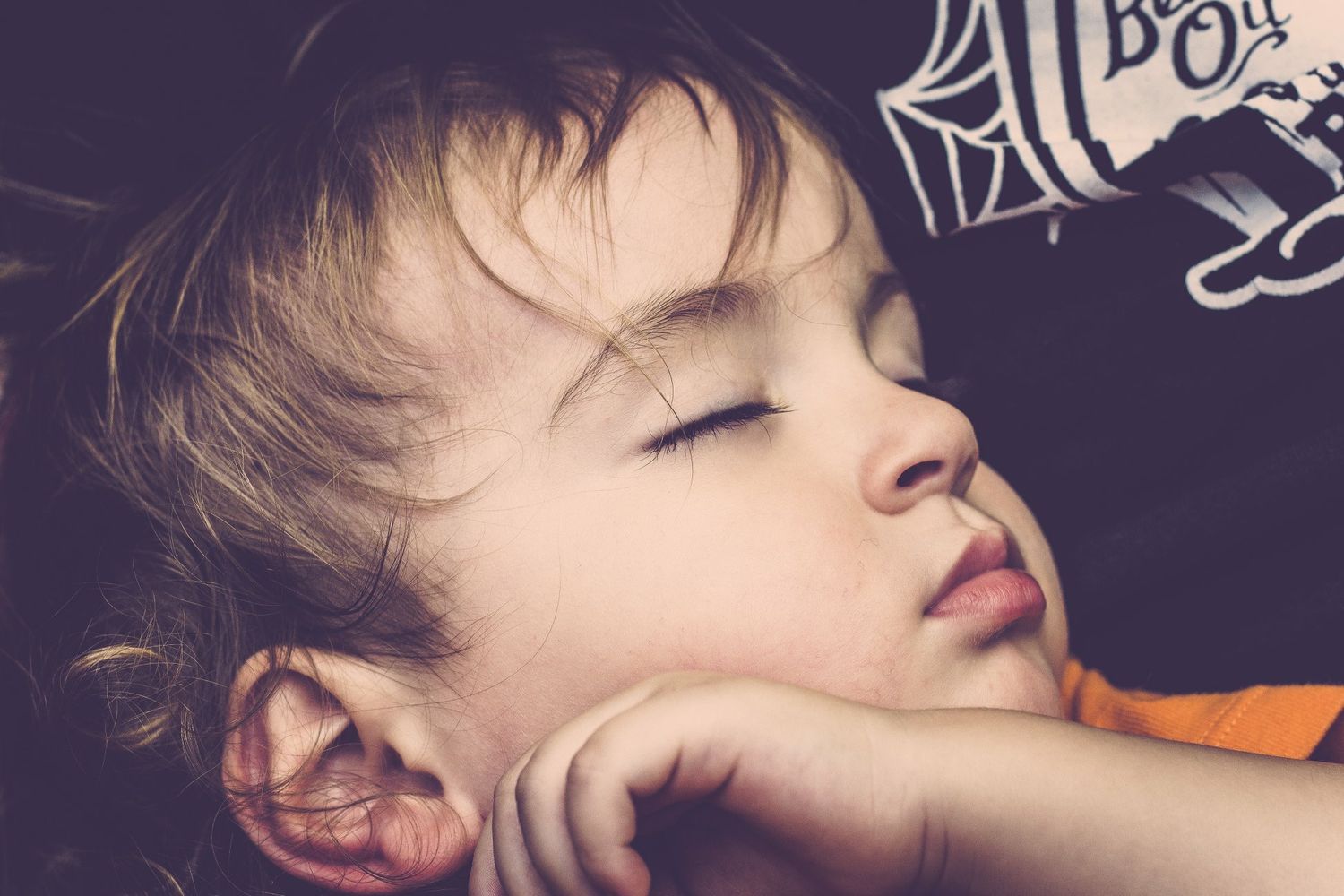 A young child sleeping peacefully with head resting on hand, wearing an orange shirt.
