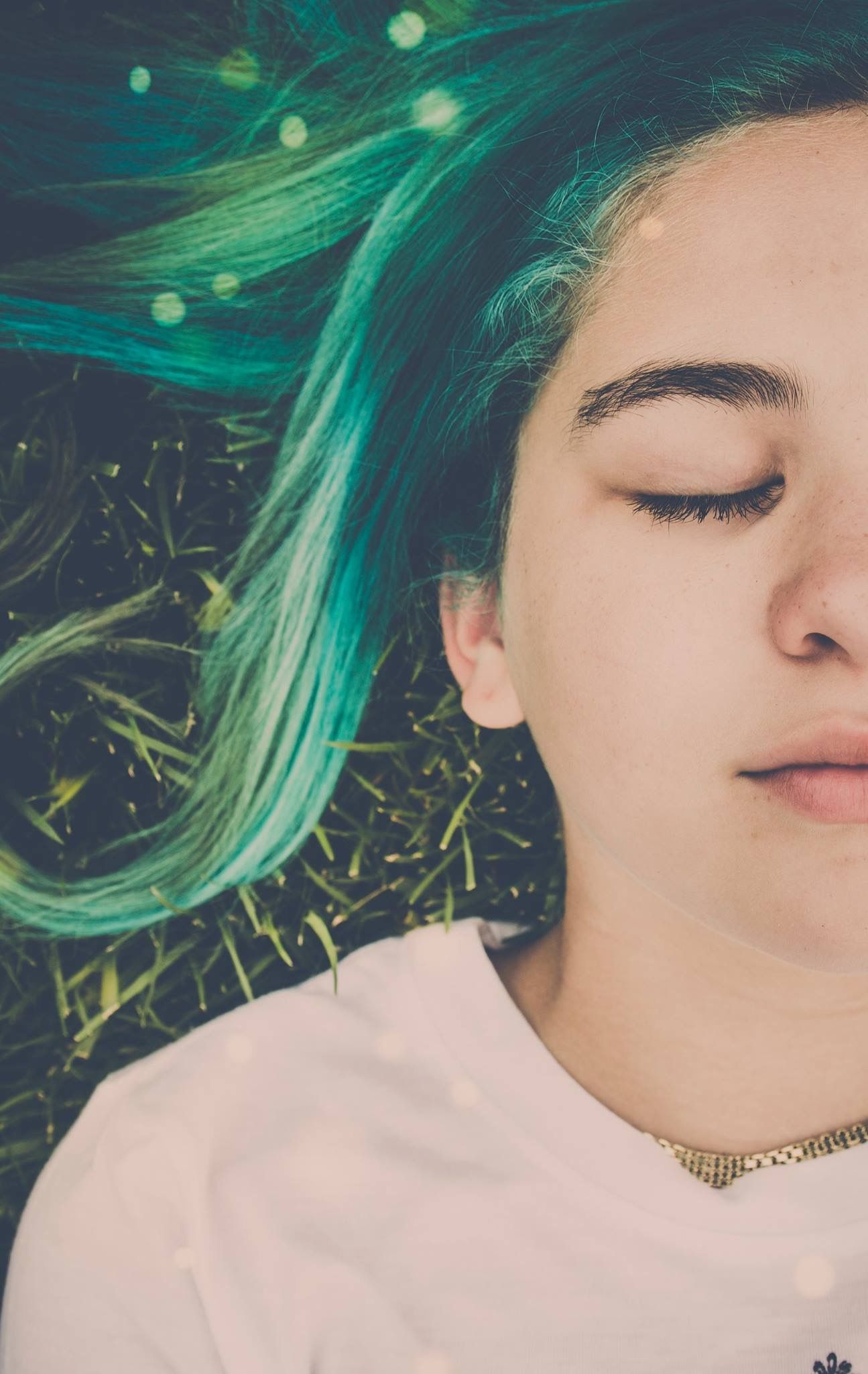 Young woman with teal and green dyed hair lying on grass with eyes closed, wearing a white top and gold necklace.
