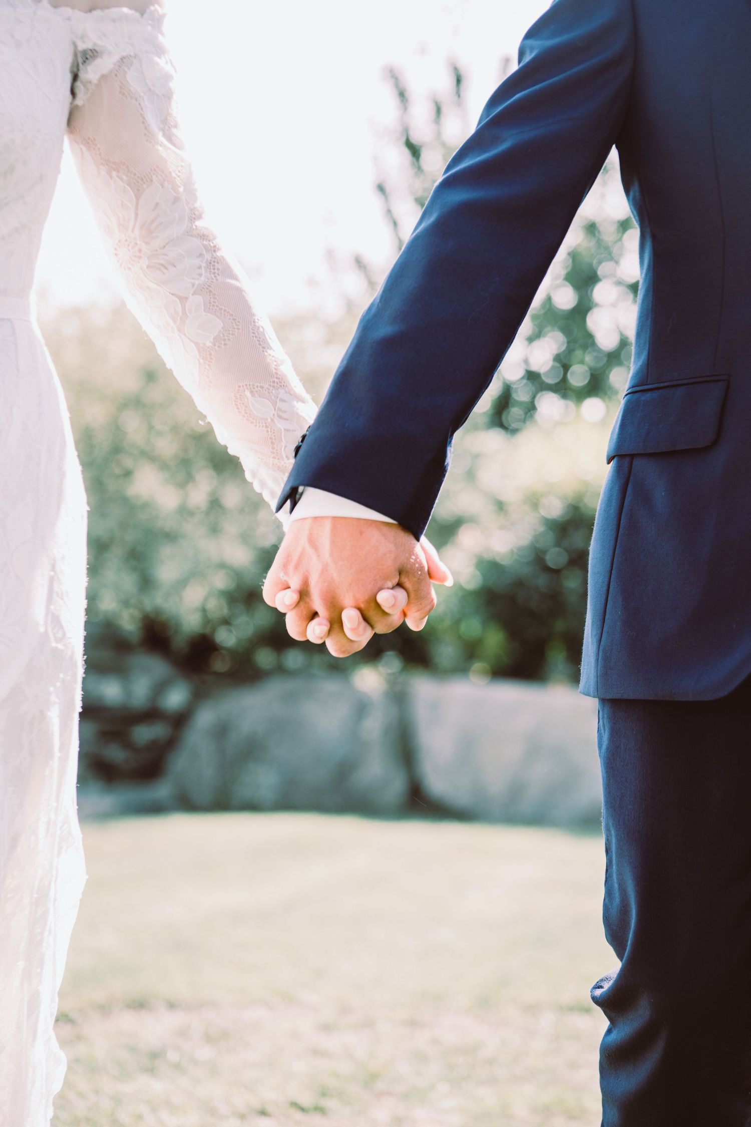 Bride and groom holding hands outdoors, showcasing wedding rings, lace dress, and navy suit in a garden setting.