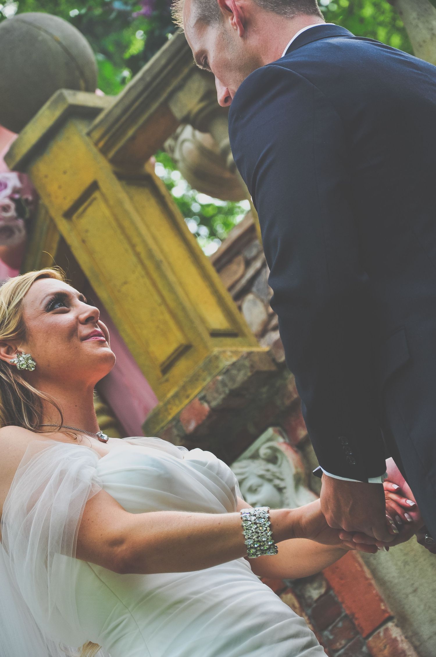 Smiling bride in white dress looks up at groom while holding hands outdoors.