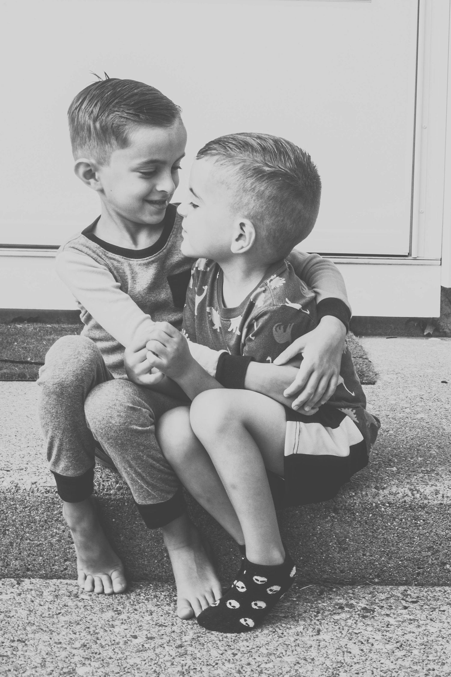 Two young boys sitting on steps, hugging and smiling at each other in a black and white photo.