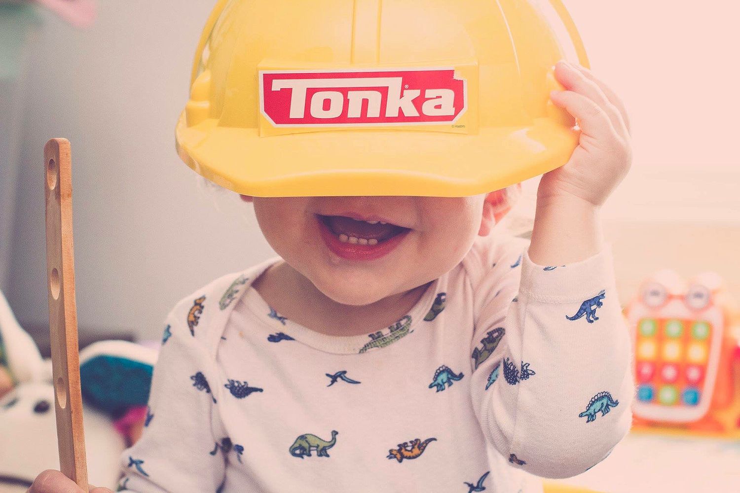 A smiling toddler wearing a yellow Tonka construction hard hat toy while dressed in a dinosaur print outfit.