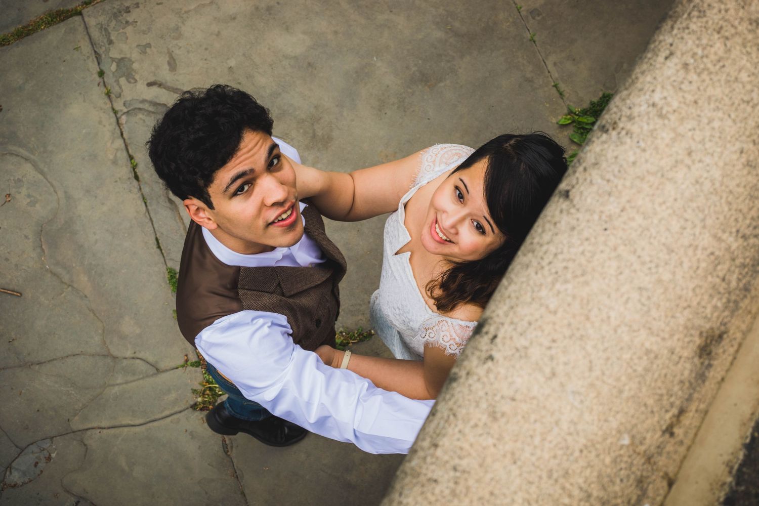Couple in wedding attire looking up at camera from below, smiling romantically near a stone wall.