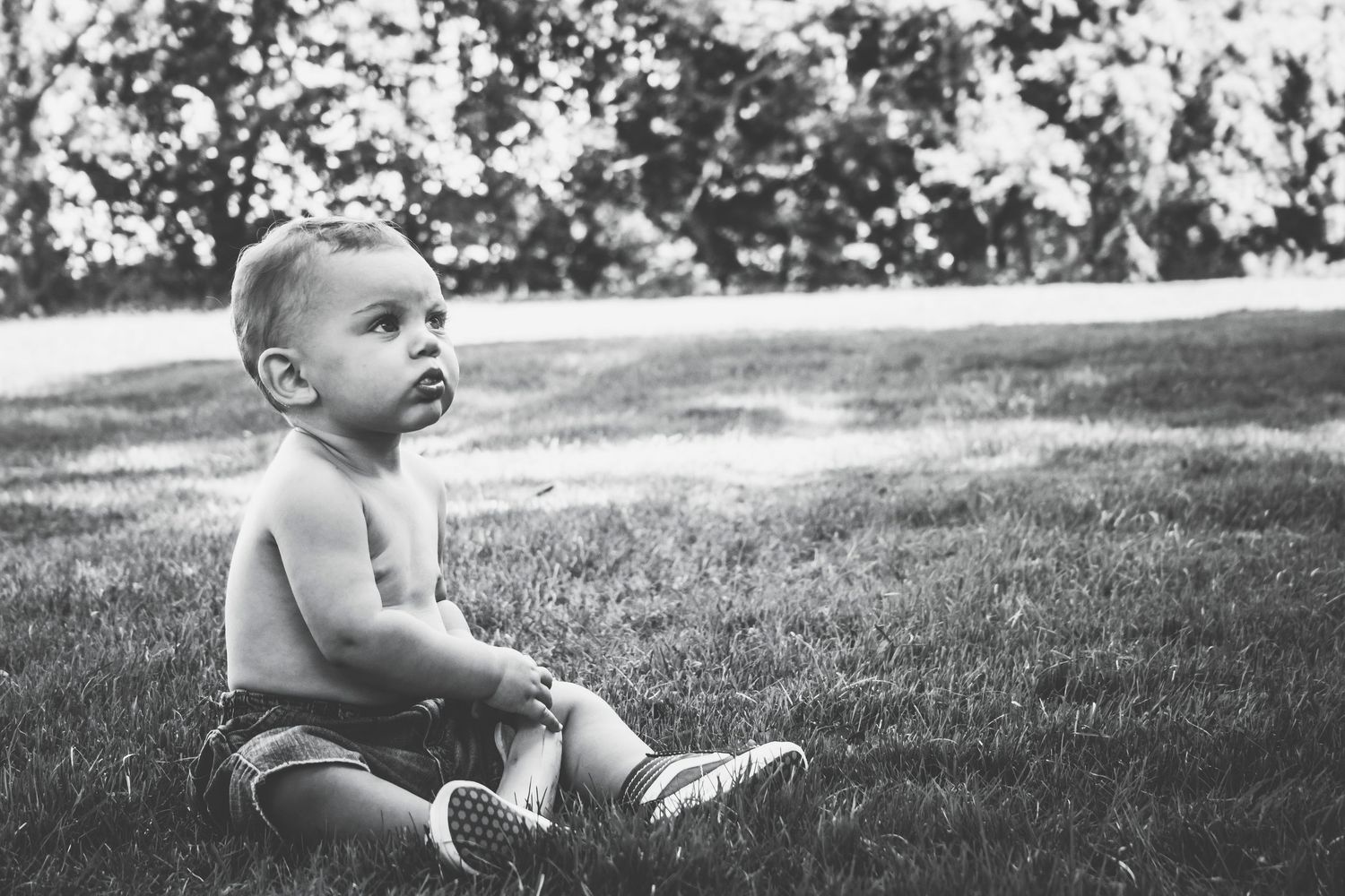 Black and white photo of a shirtless toddler sitting on green grass in a park, wearing shorts and sneakers.