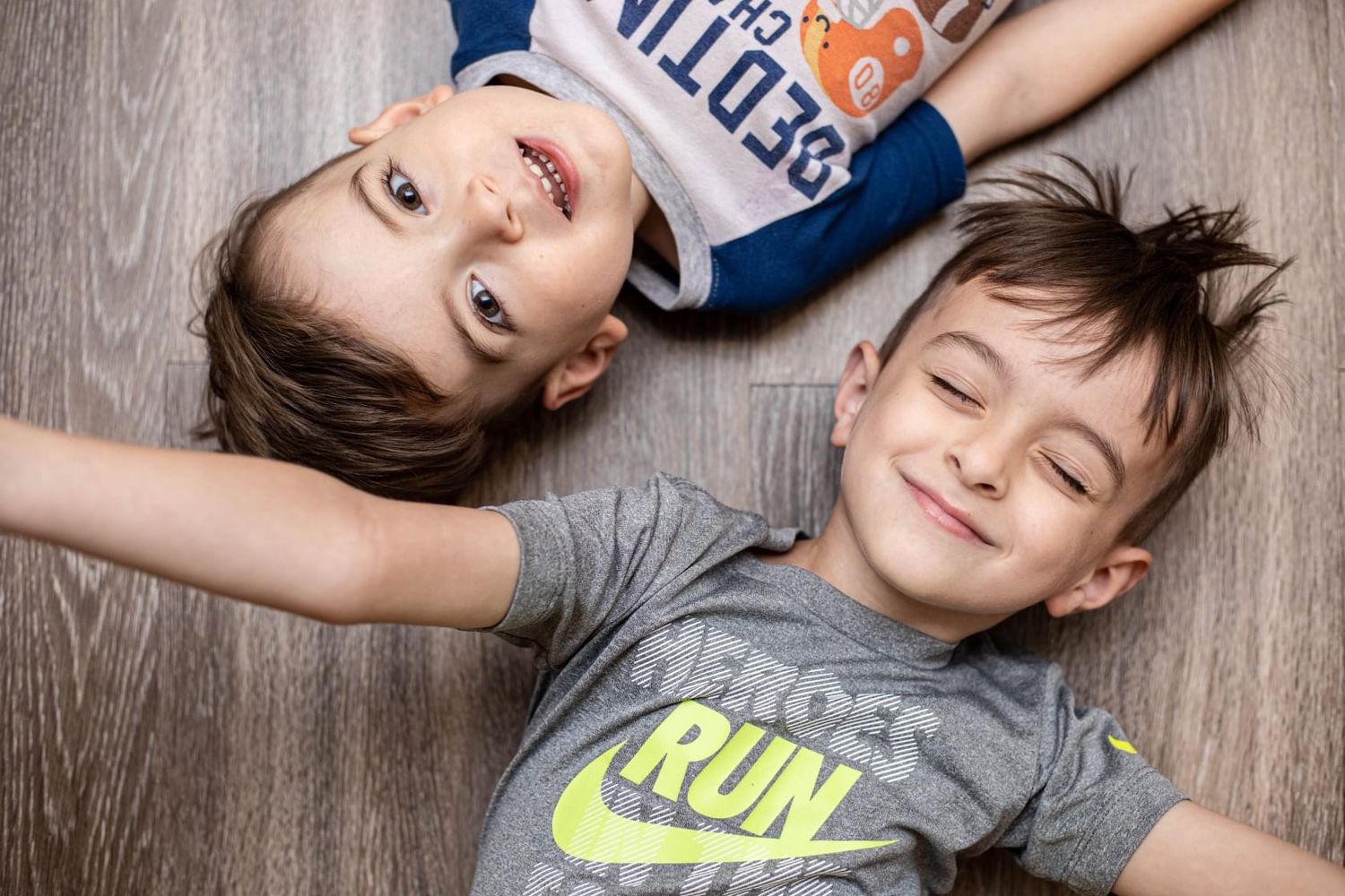 Two happy boys lying on a wooden floor, one wearing a Nike shirt, smiling and posing for a top-down photo.