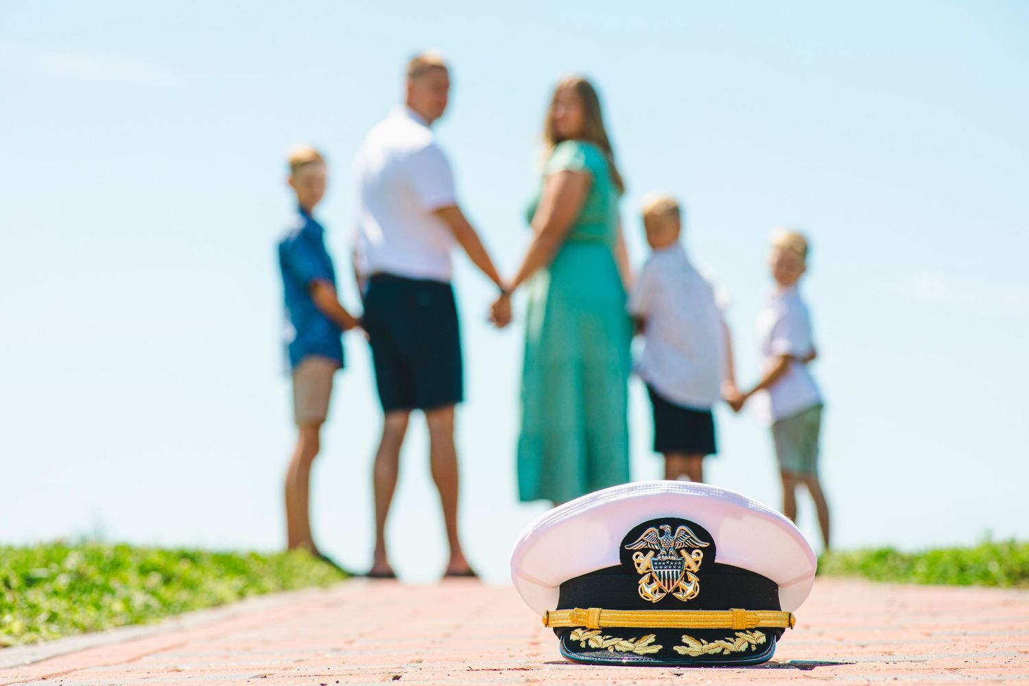 Navy officer cap in foreground with military family holding hands on sunny brick path outdoors.