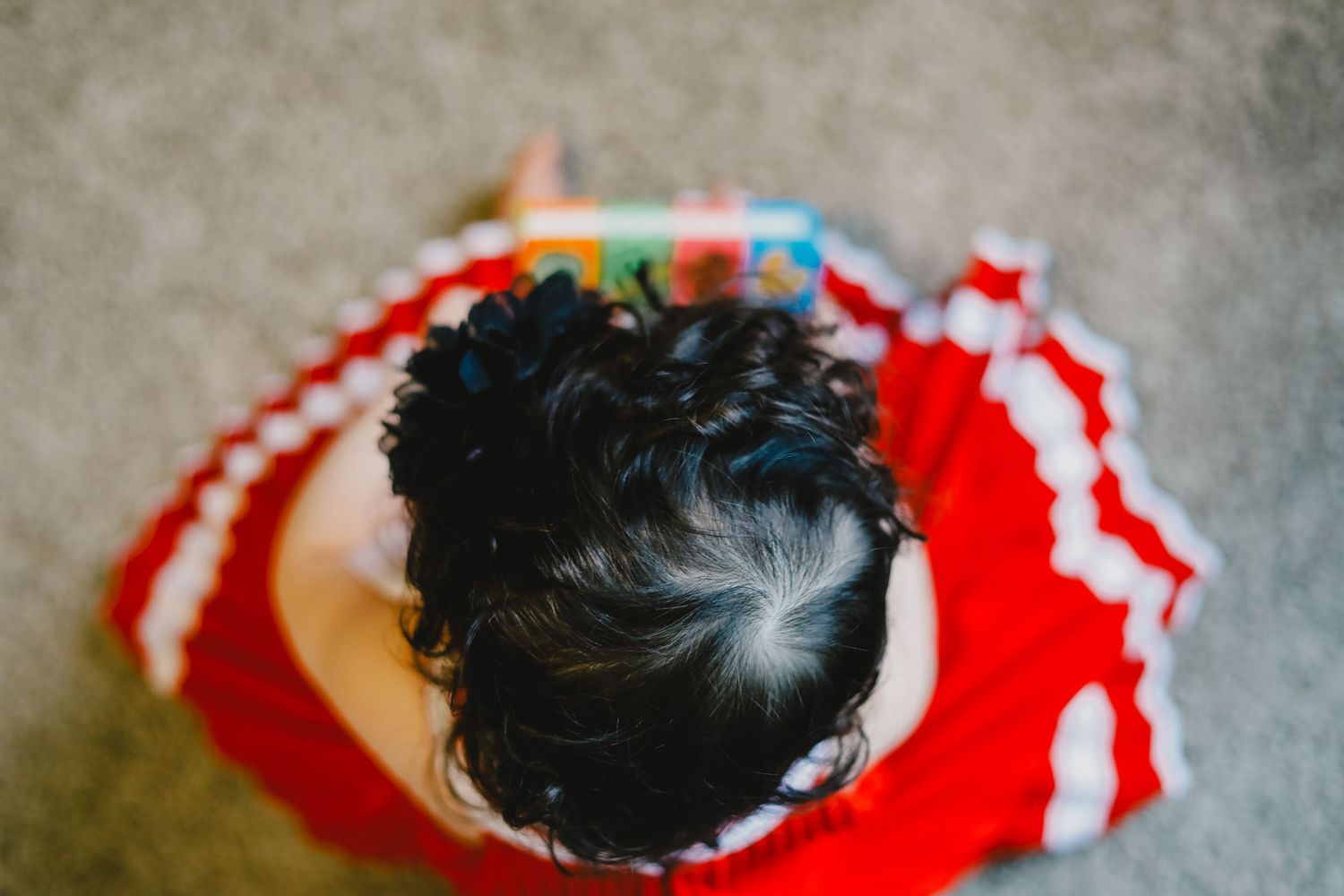 Overhead view of a young child with dark curly hair in a red dress reading a colorful book on the floor.