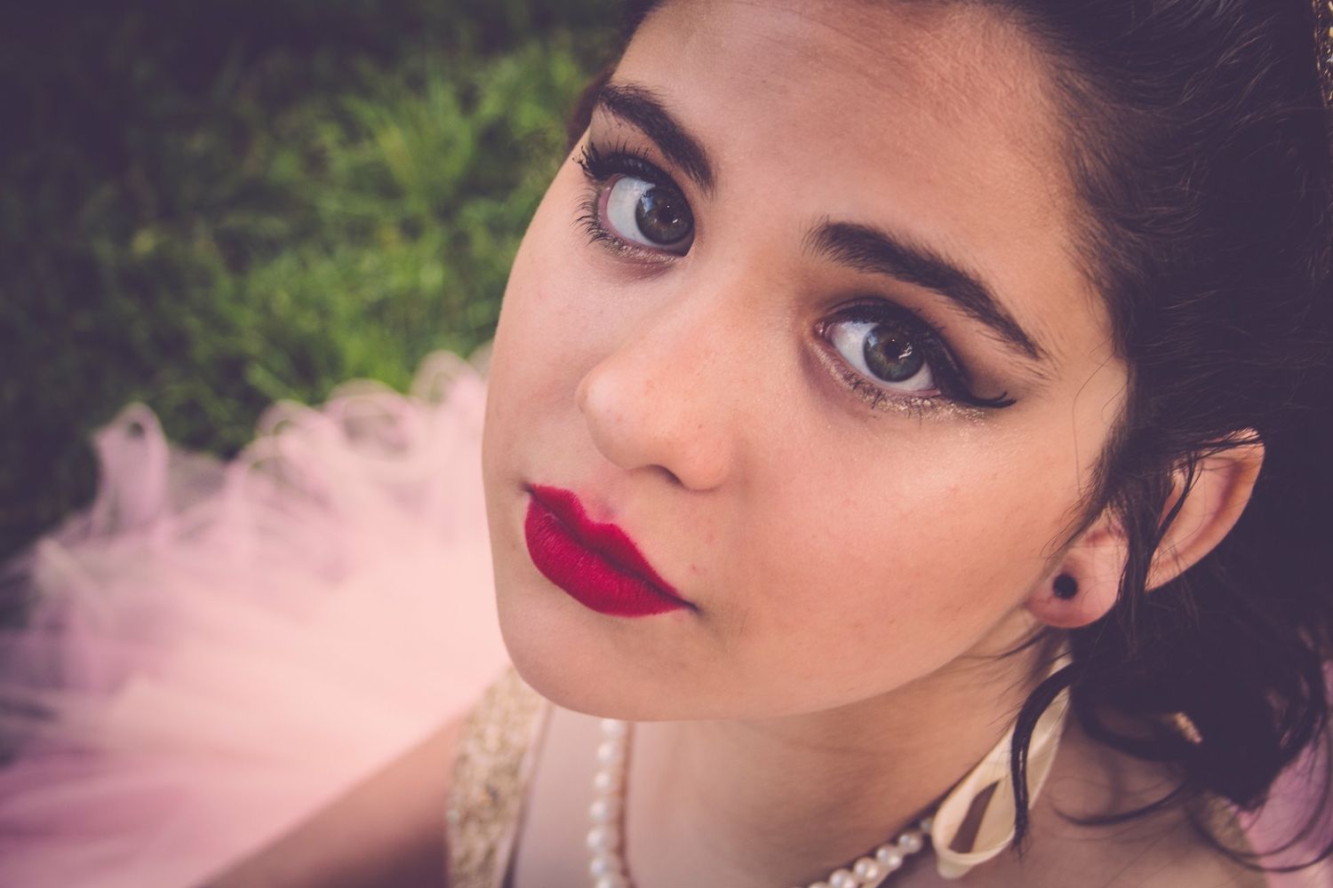 Young woman with bold red lips, dramatic eye makeup, and pearl necklace wearing a pink tulle dress outdoors.