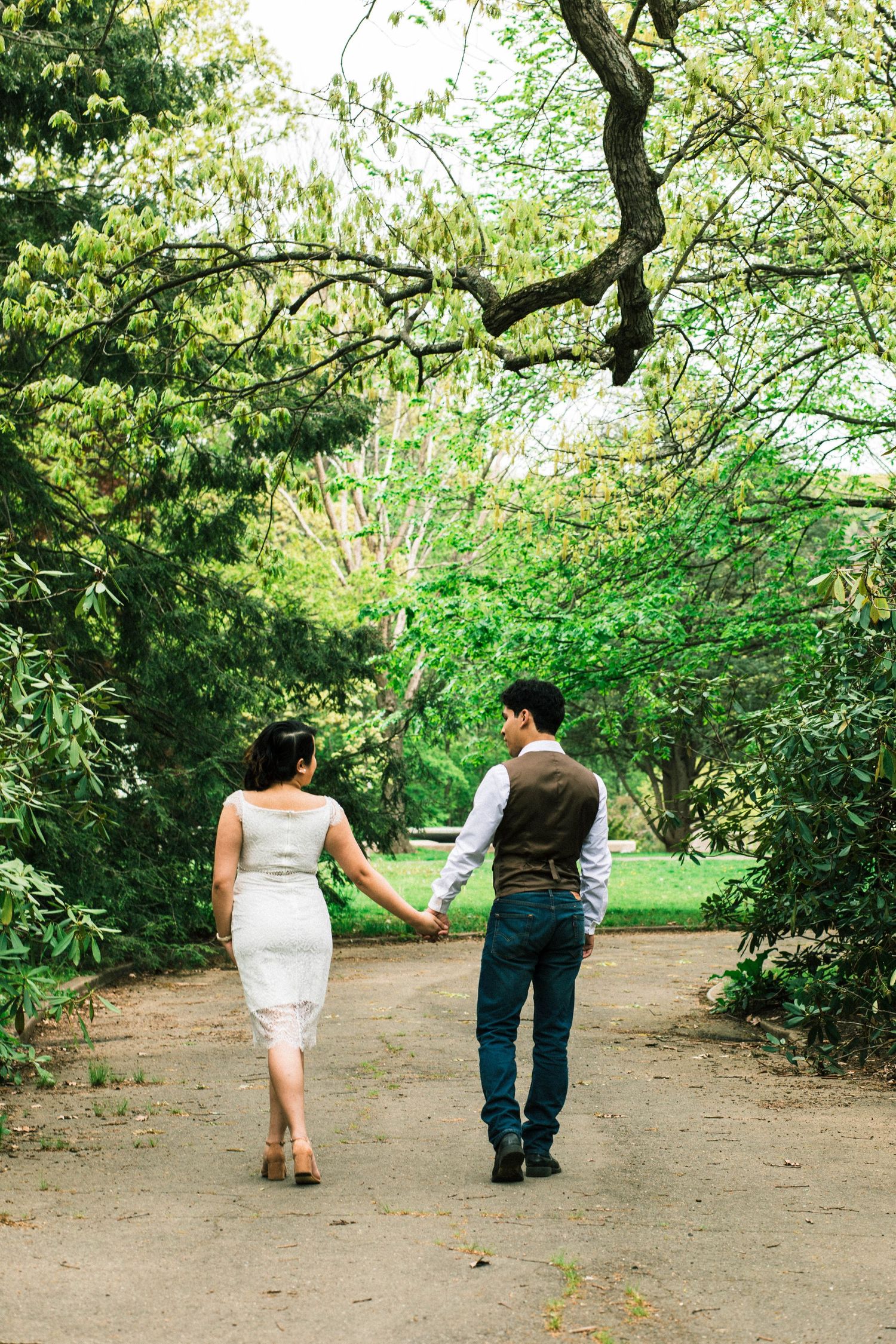 Bride and groom walking hand in hand down a tree-lined garden path, viewed from behind.