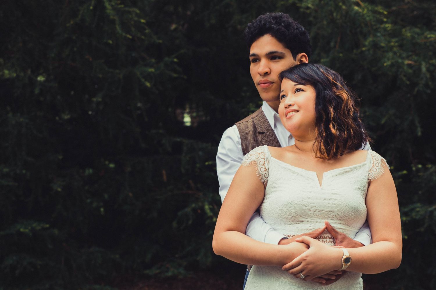 A couple in wedding attire embraces outdoors, the groom holding the bride from behind among lush green trees.