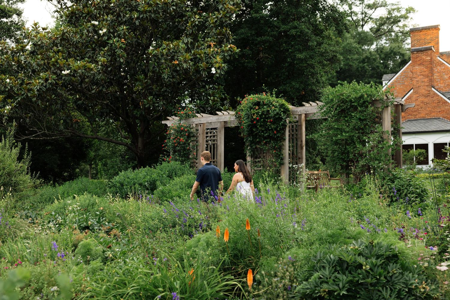 Two people walking through a lush garden path toward a wooden pergola surrounded by wildflowers and greenery.