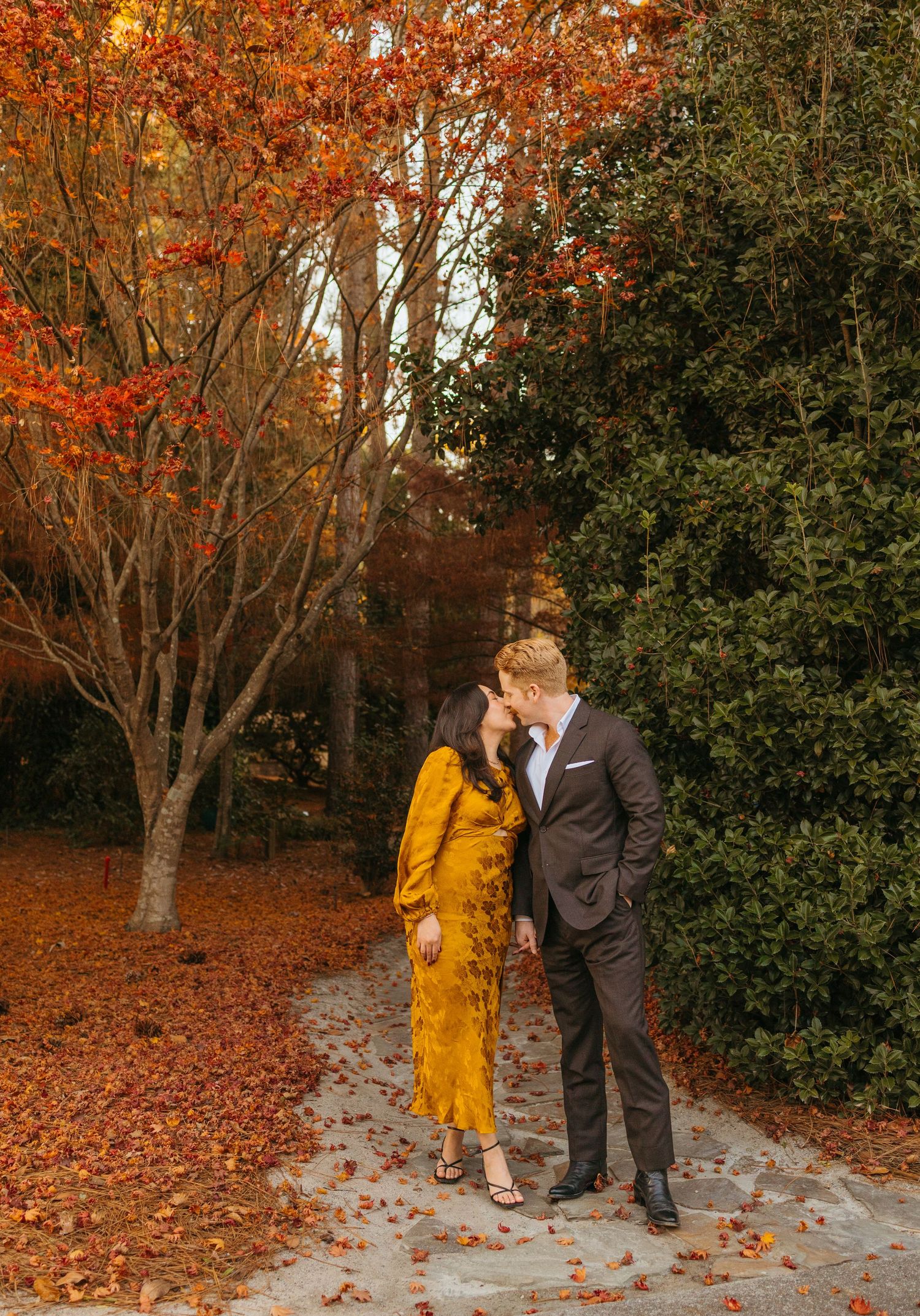 Couple sharing a kiss on a leaf-strewn path surrounded by stunning red autumn foliage and green hedges.