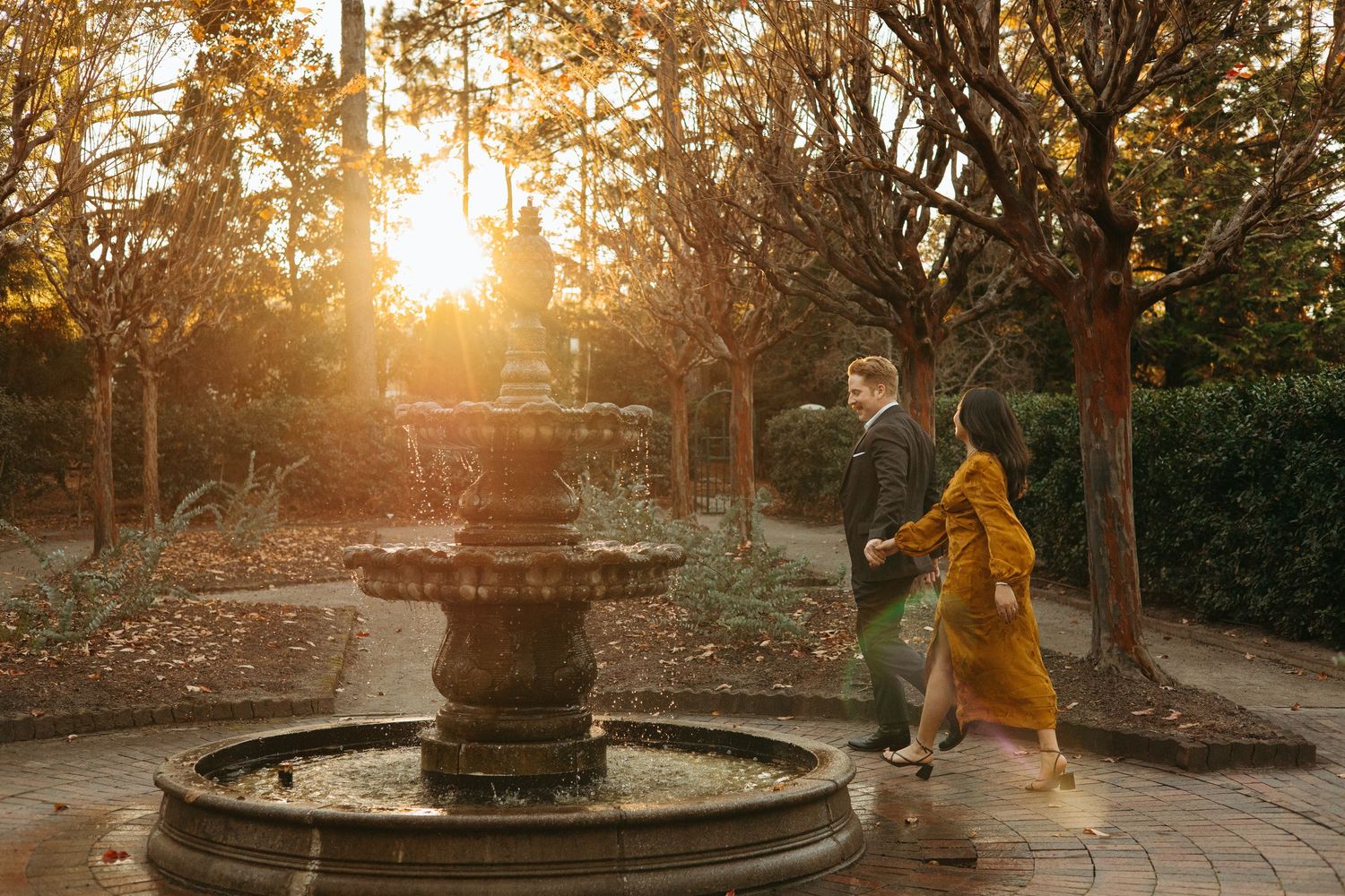 Couple walking past an ornate tiered fountain in a garden bathed in warm golden sunset light.