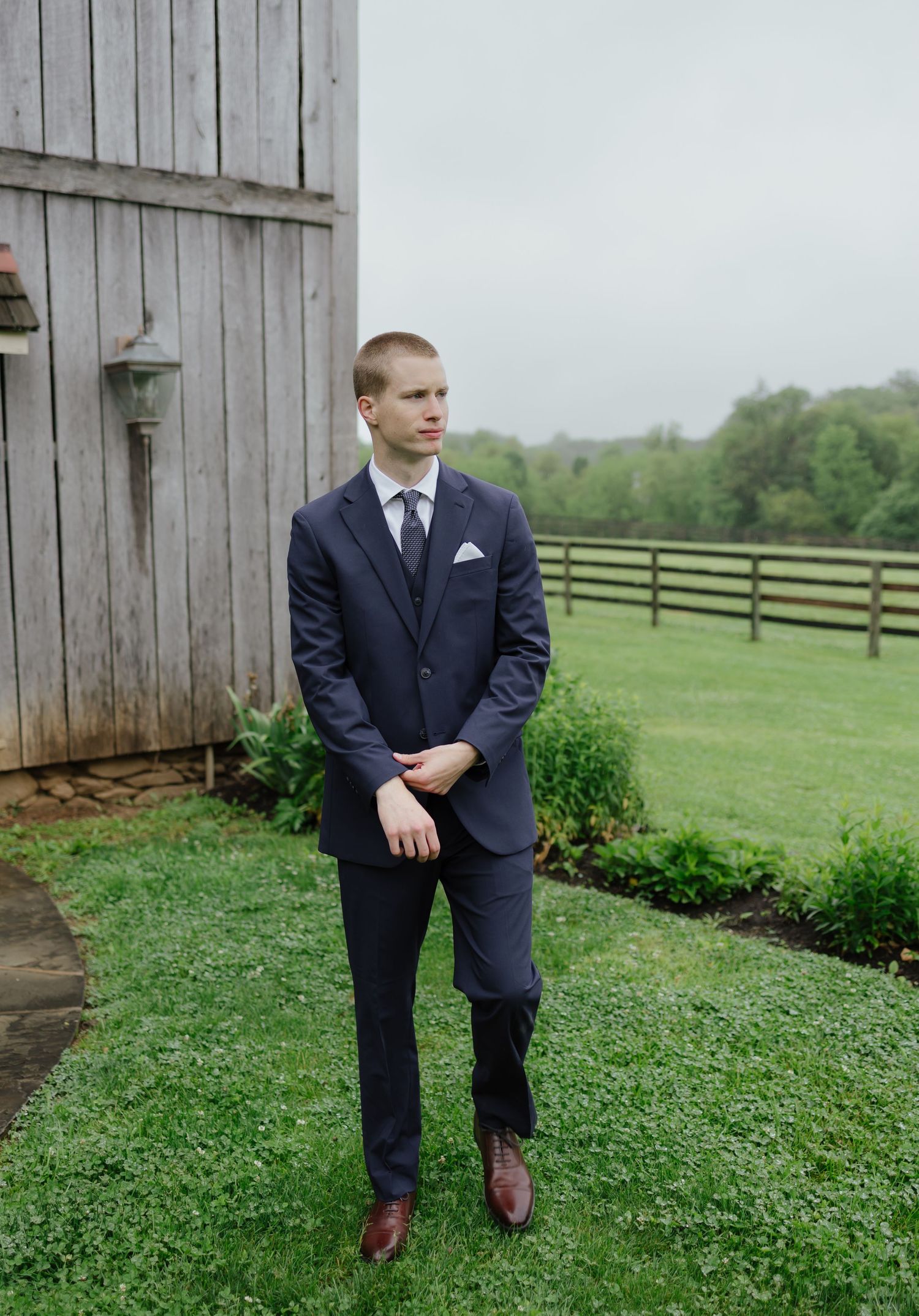 Groom in navy three-piece suit with white pocket square stands on green lawn beside rustic barn.