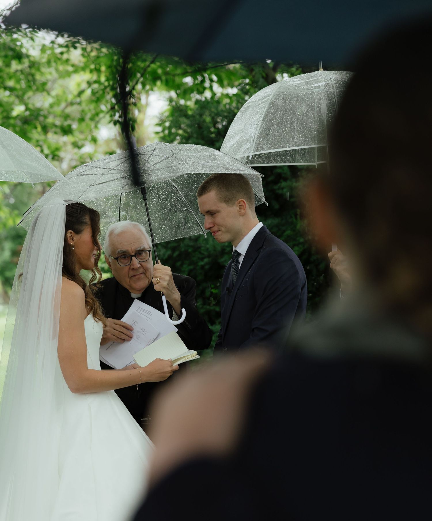 Bride reads vows to groom during intimate outdoor wedding ceremony under clear umbrellas.