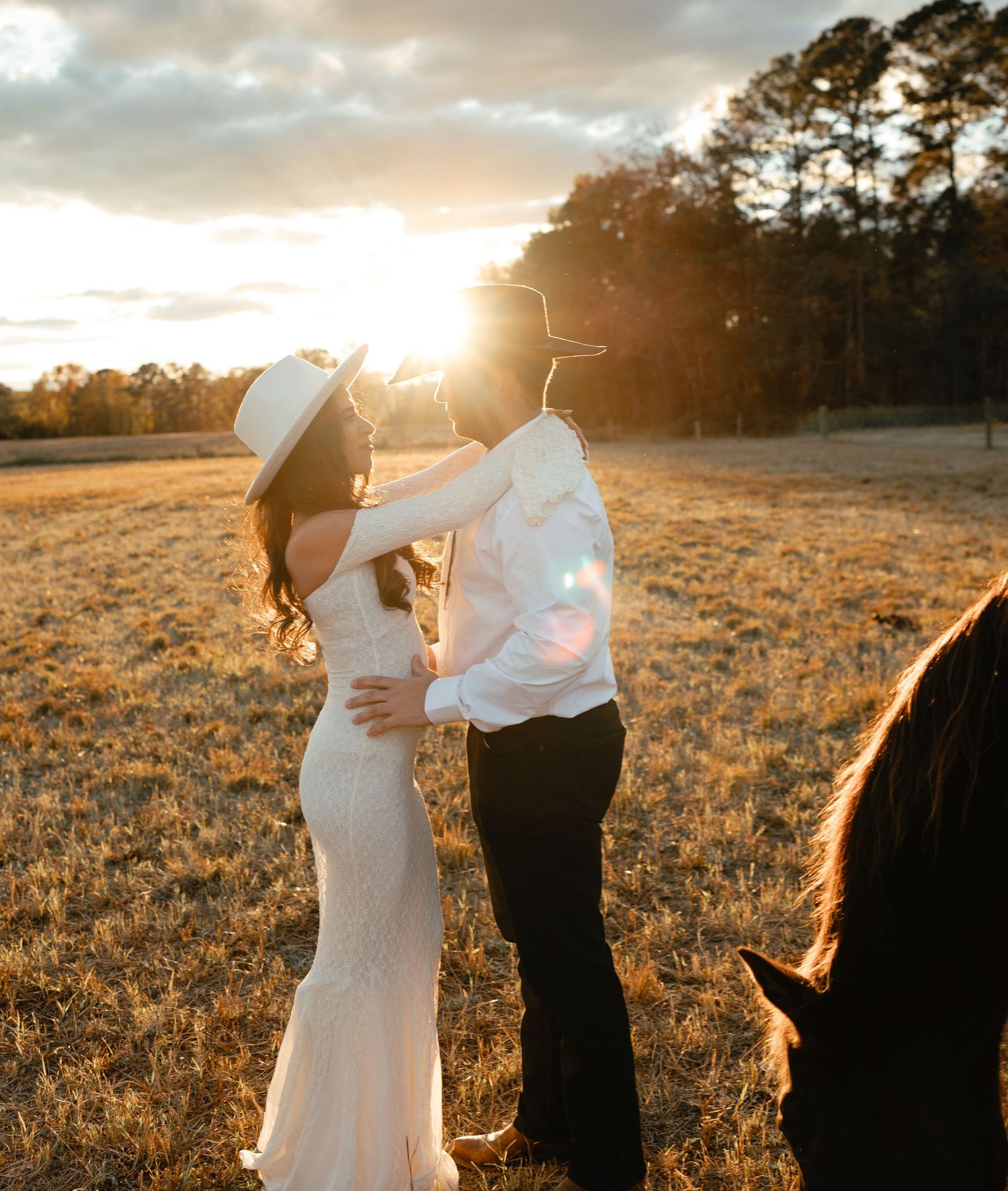 Couple sharing a romantic moment at golden hour in a field, bride in white dress and groom in cowboy hat.