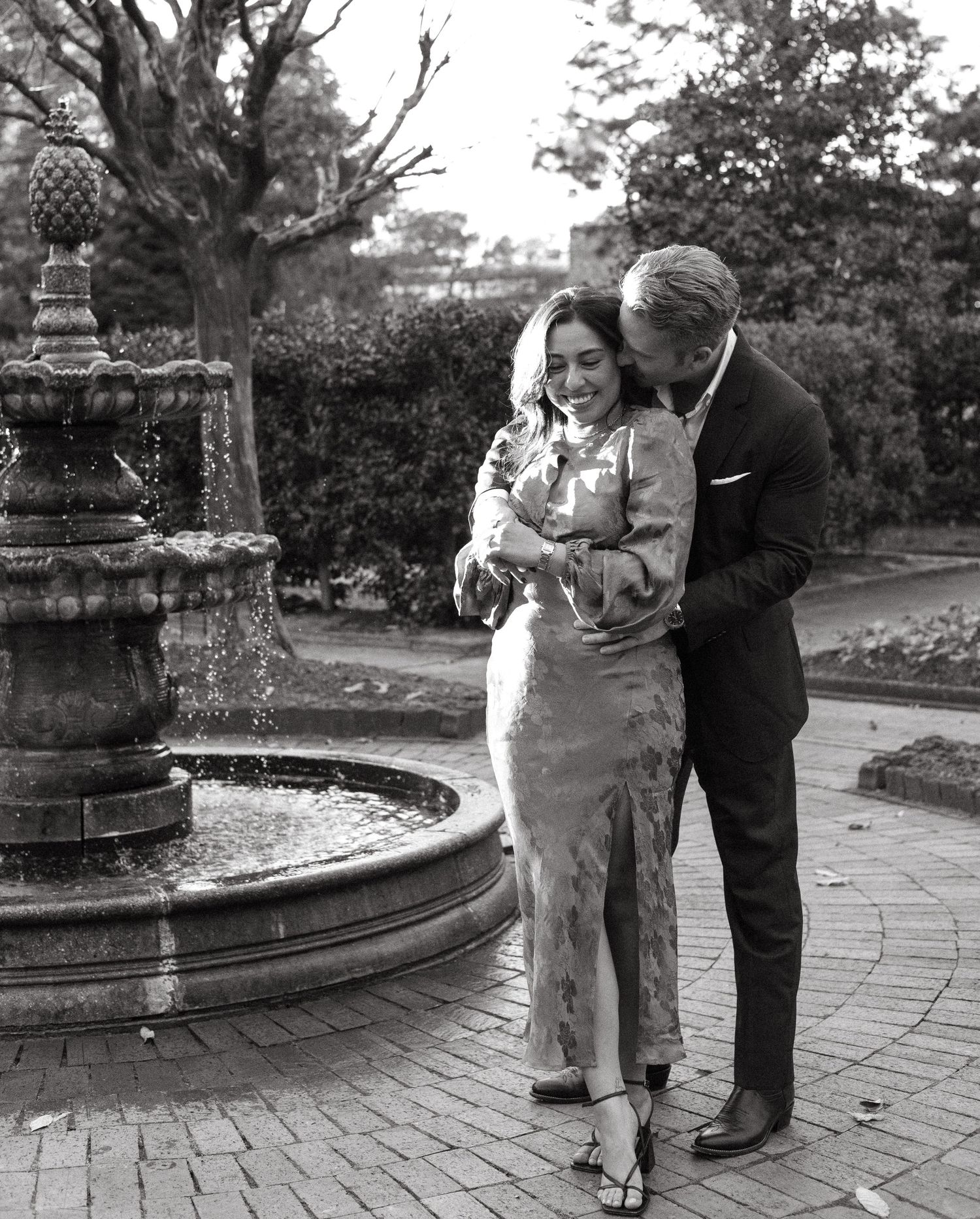 Black and white photo of a joyful couple embracing and laughing beside a garden fountain.