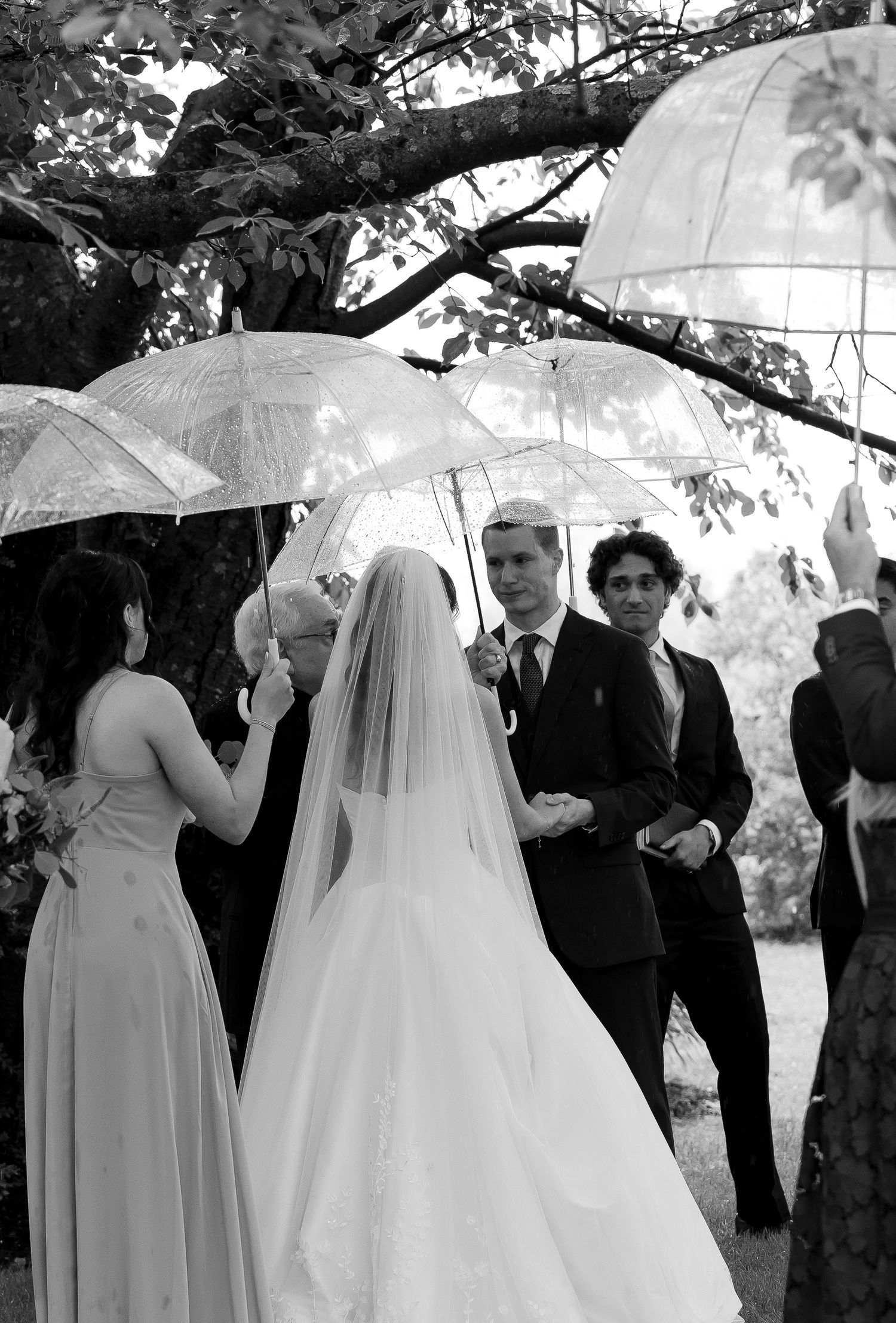 Black and white photo of bride in veil and wedding party holding clear umbrellas during outdoor rainy ceremony.
