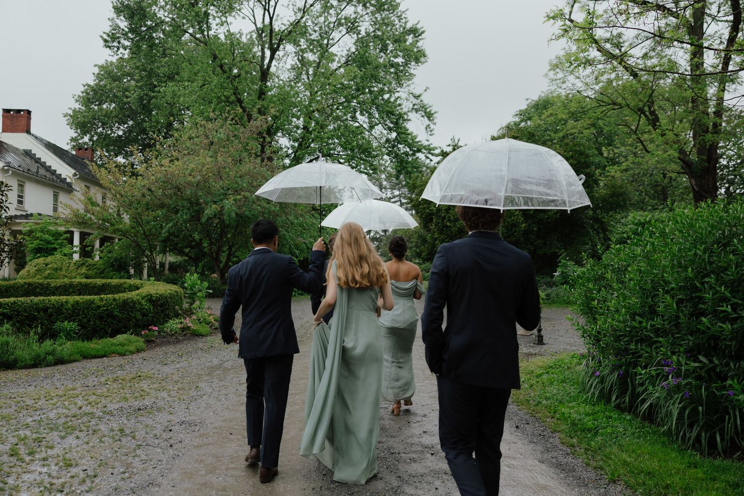 Wedding party walking down a garden path with clear bubble umbrellas on a rainy day.
