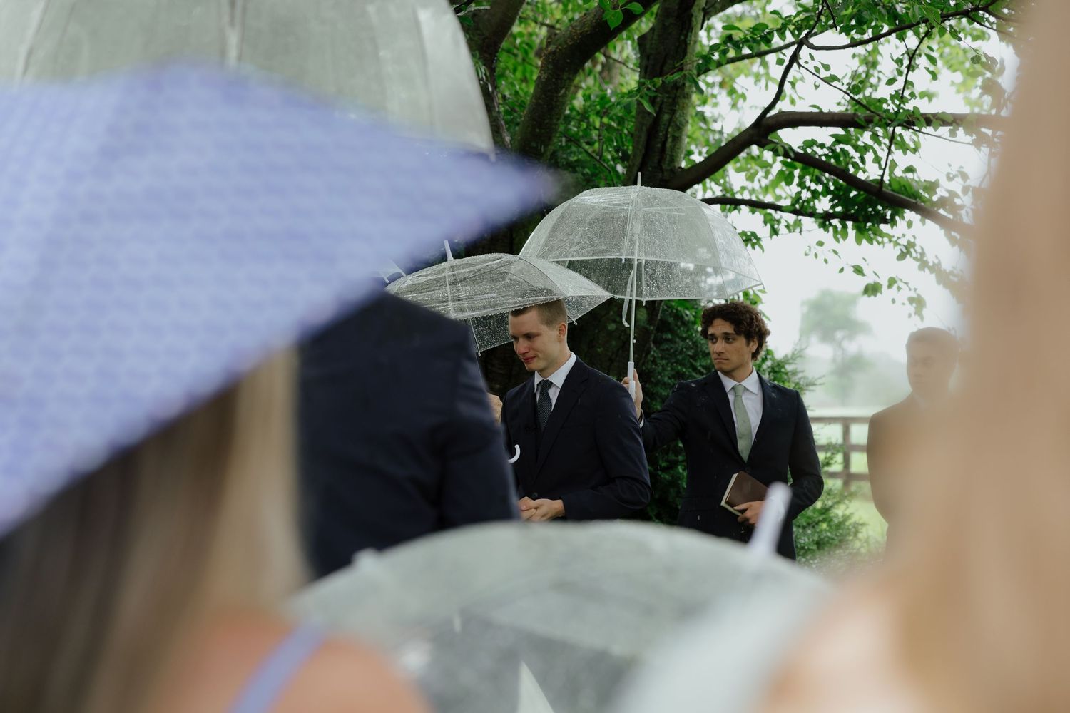 Groomsmen holding clear bubble umbrellas during an outdoor rainy wedding ceremony.