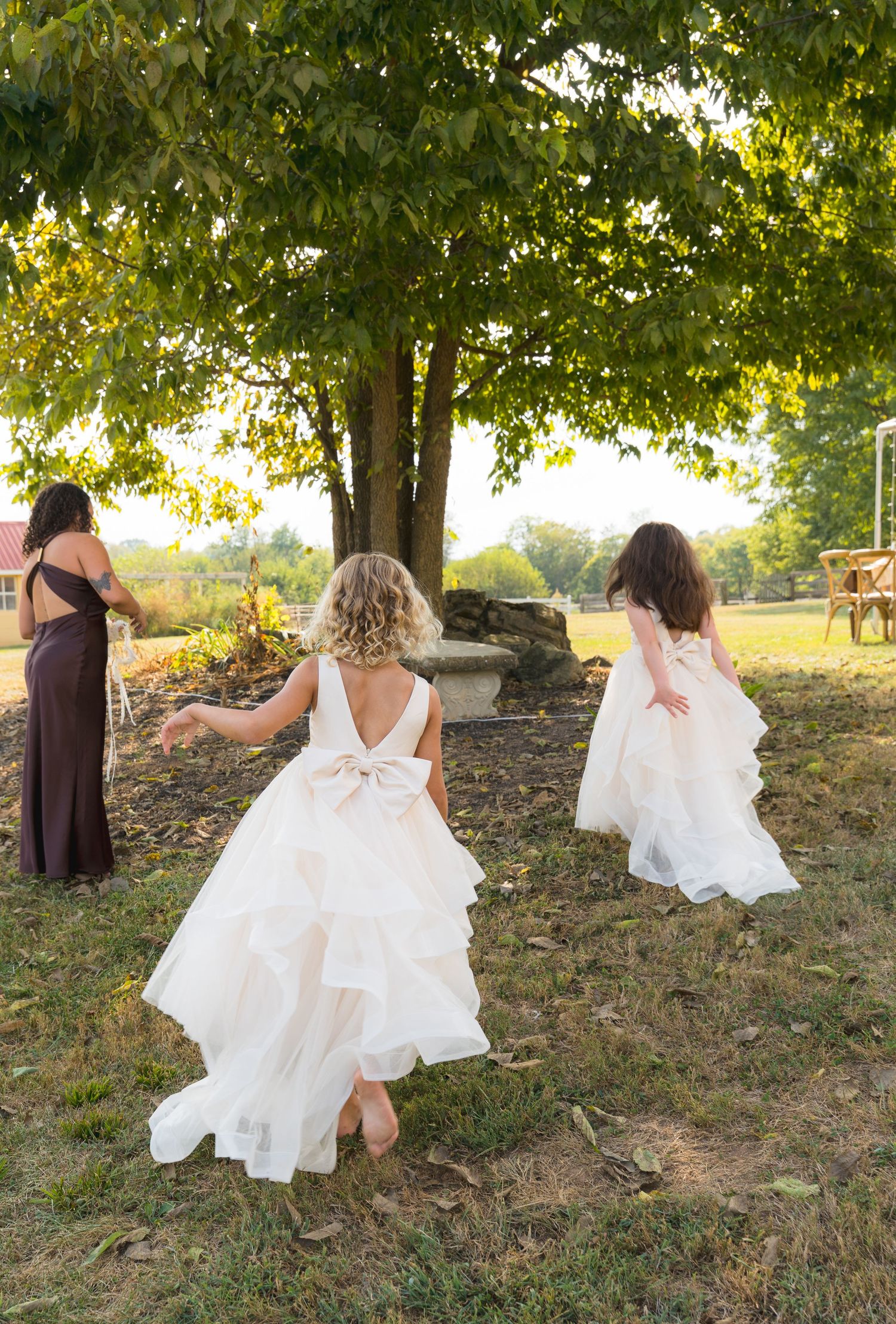 Two flower girls in white ruffled dresses running barefoot across green lawn at outdoor wedding.