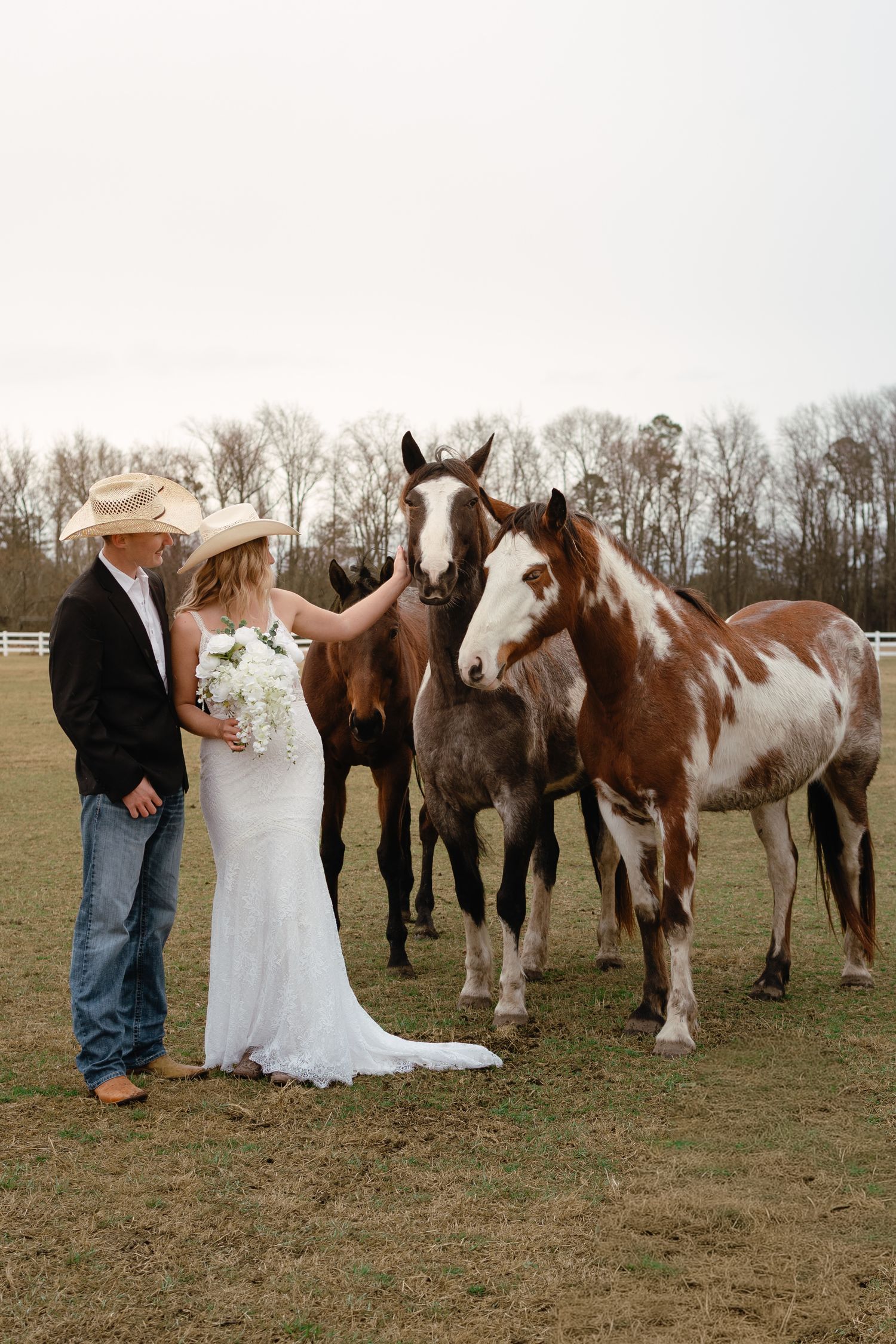 Bride and groom in cowboy hats posing with paint horses on a farm in wedding attire.