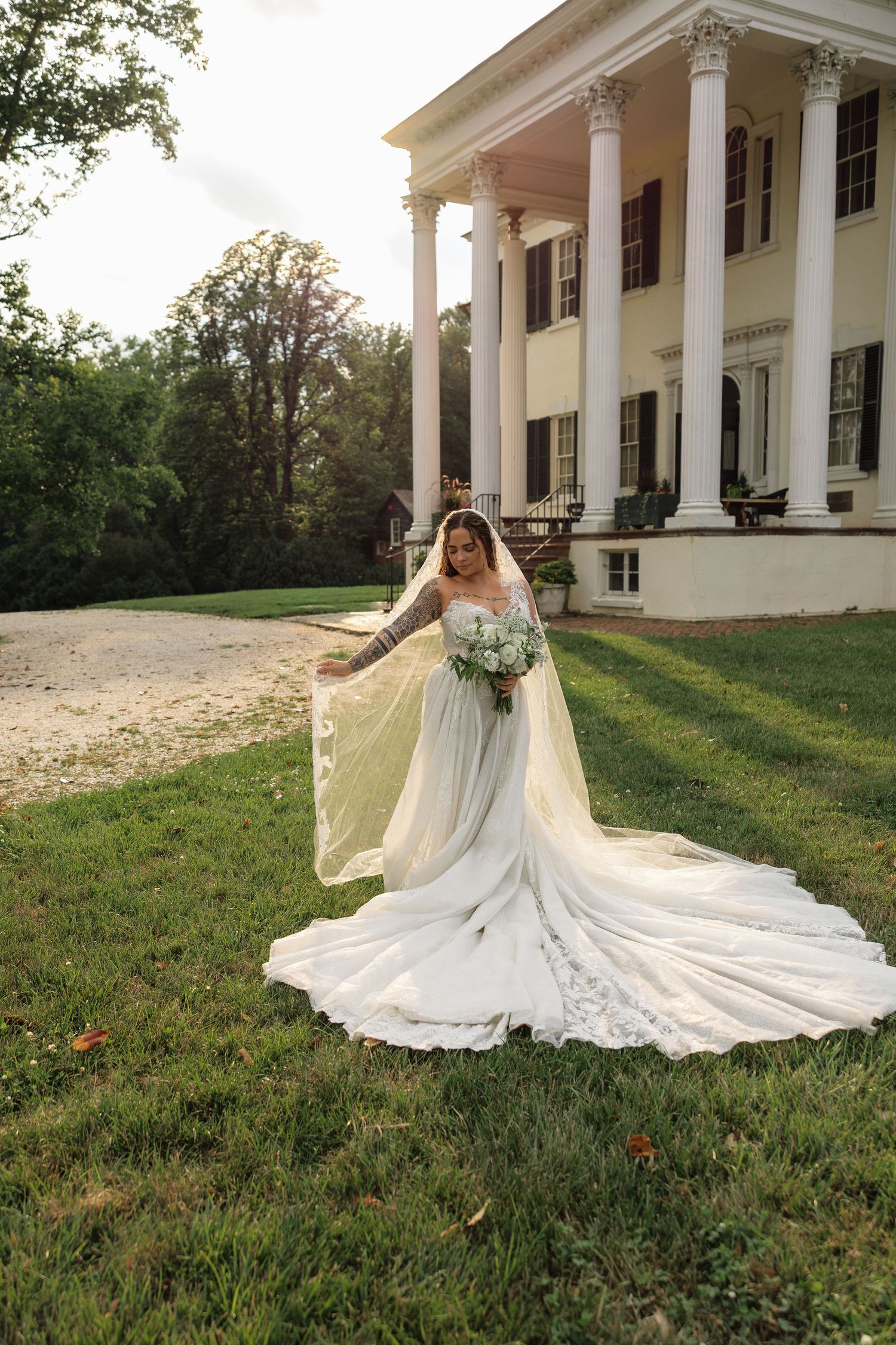 Bride in a flowing white lace gown with veil poses on green lawn before a grand white colonial mansion.