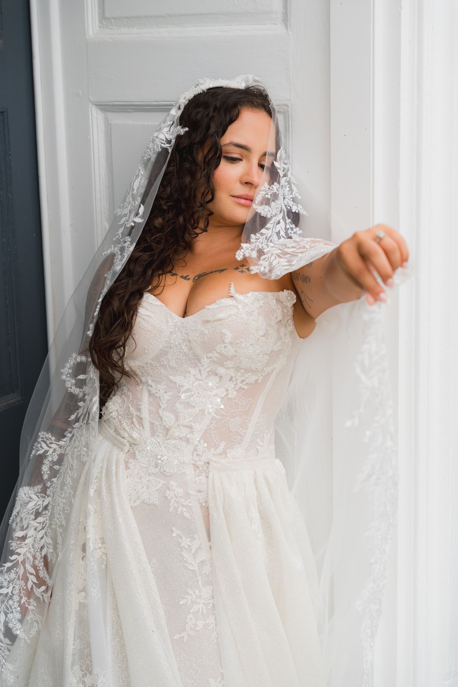 Bride in lace wedding gown and veil with curly hair posing elegantly by white door indoors.
