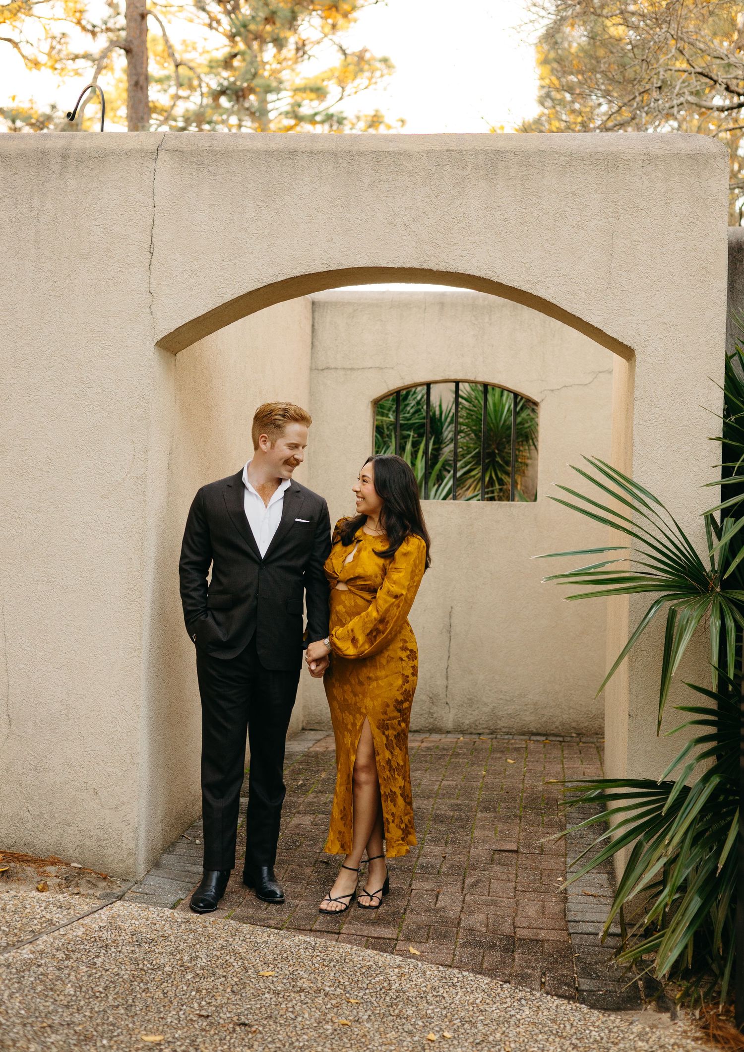 Couple holding hands under a stone archway, man in black suit and woman in mustard yellow dress, smiling at each other.