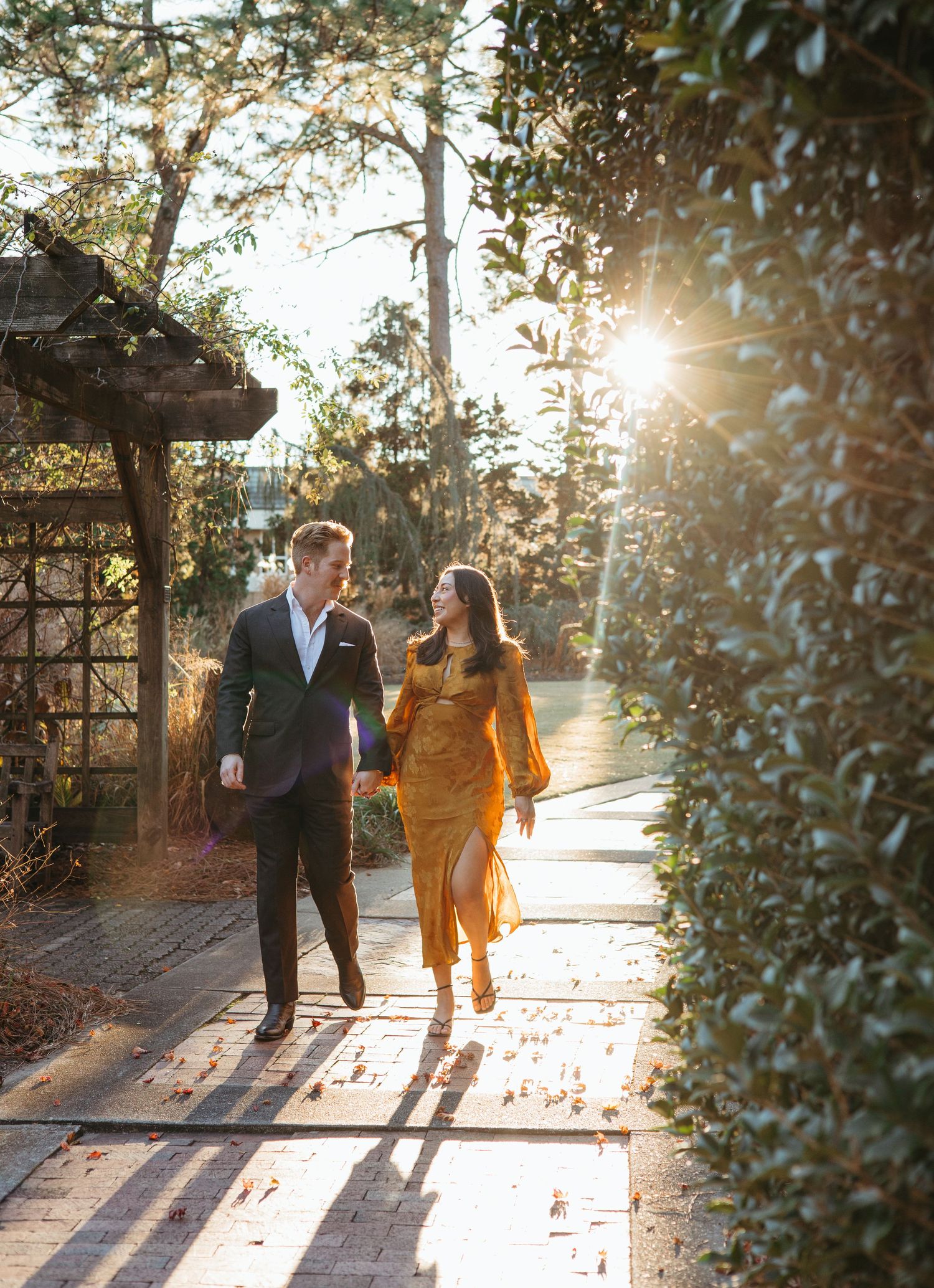 Couple strolling along a sun-drenched garden walkway surrounded by lush hedges and warm light.