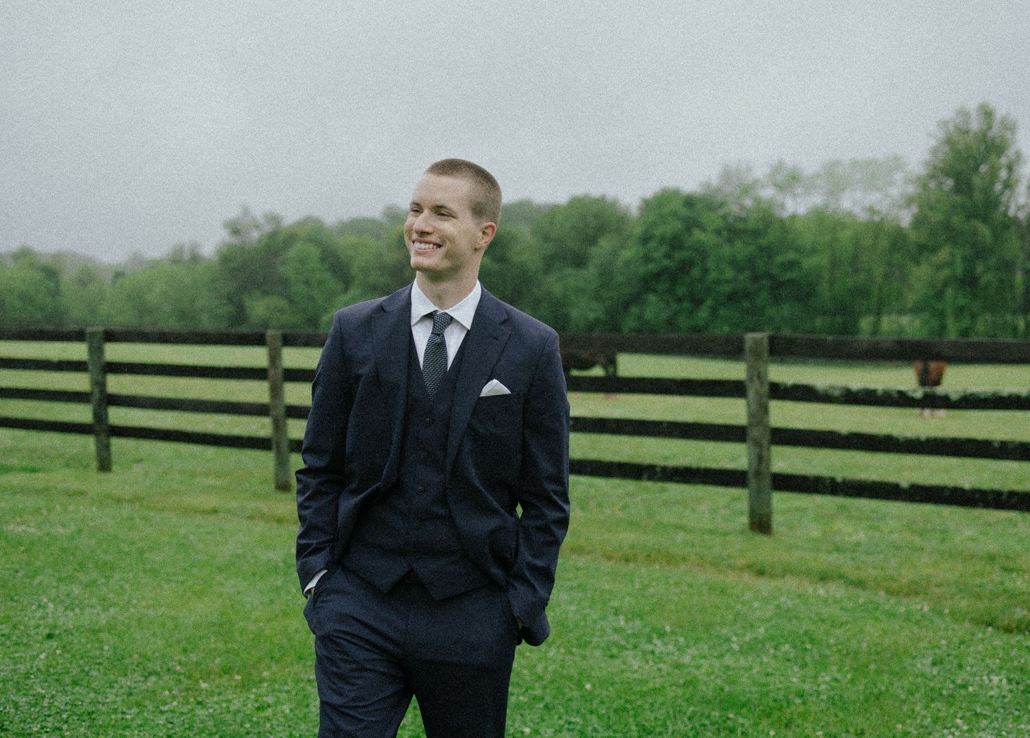 Smiling groomsman in navy three-piece suit standing on green grass with fence backdrop.