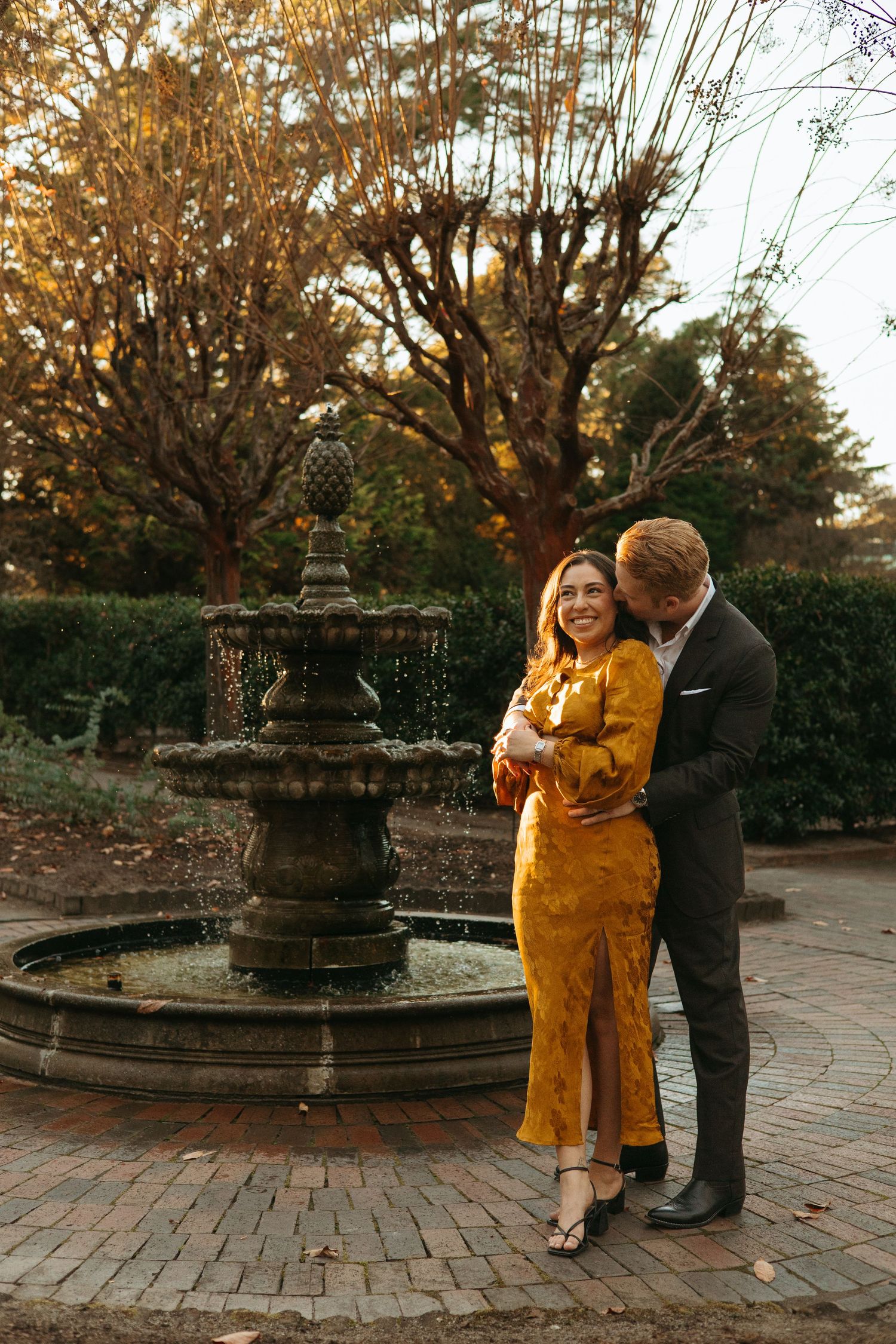 Couple laughing together beside a tiered stone fountain in an autumn garden at sunset.